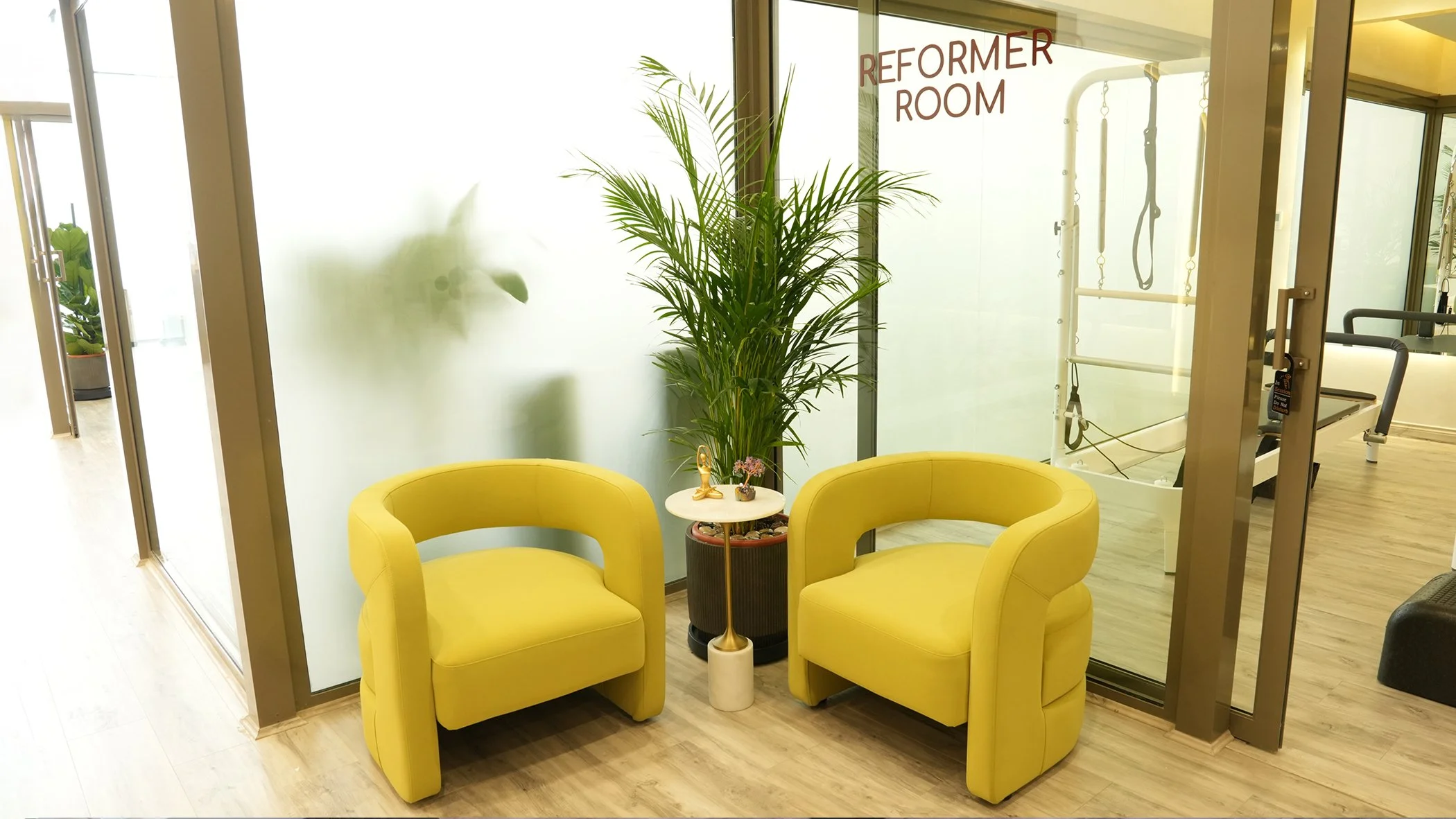 Two yellow armchairs with a small white table between them, a large potted plant behind the table, and frosted glass glass wall with red lettering reading 'Reformer Room' on it.
