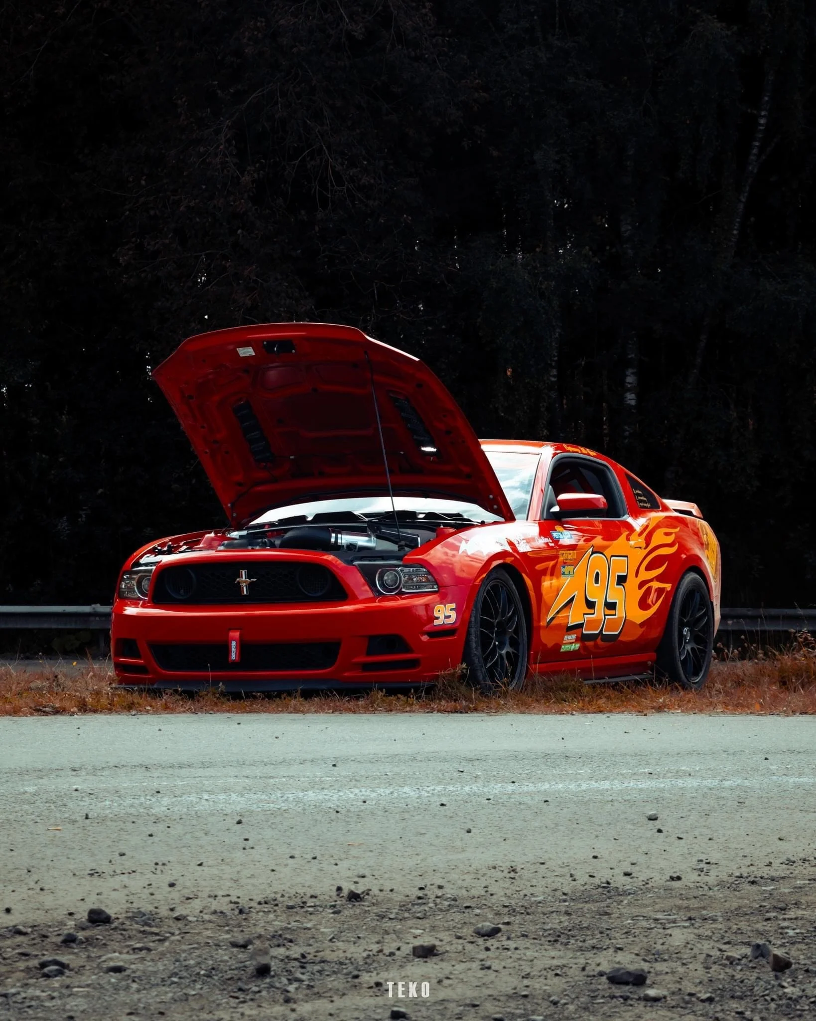 Voiture de course rouge avec le décalque du numéro 95 et des flammes, stationnée avec le capot ouvert sur le bord de la route dans une zone boisée .
La voiture est une Ford Mustang 5.0 au couleur de Flash Mcqueen du film Cars .