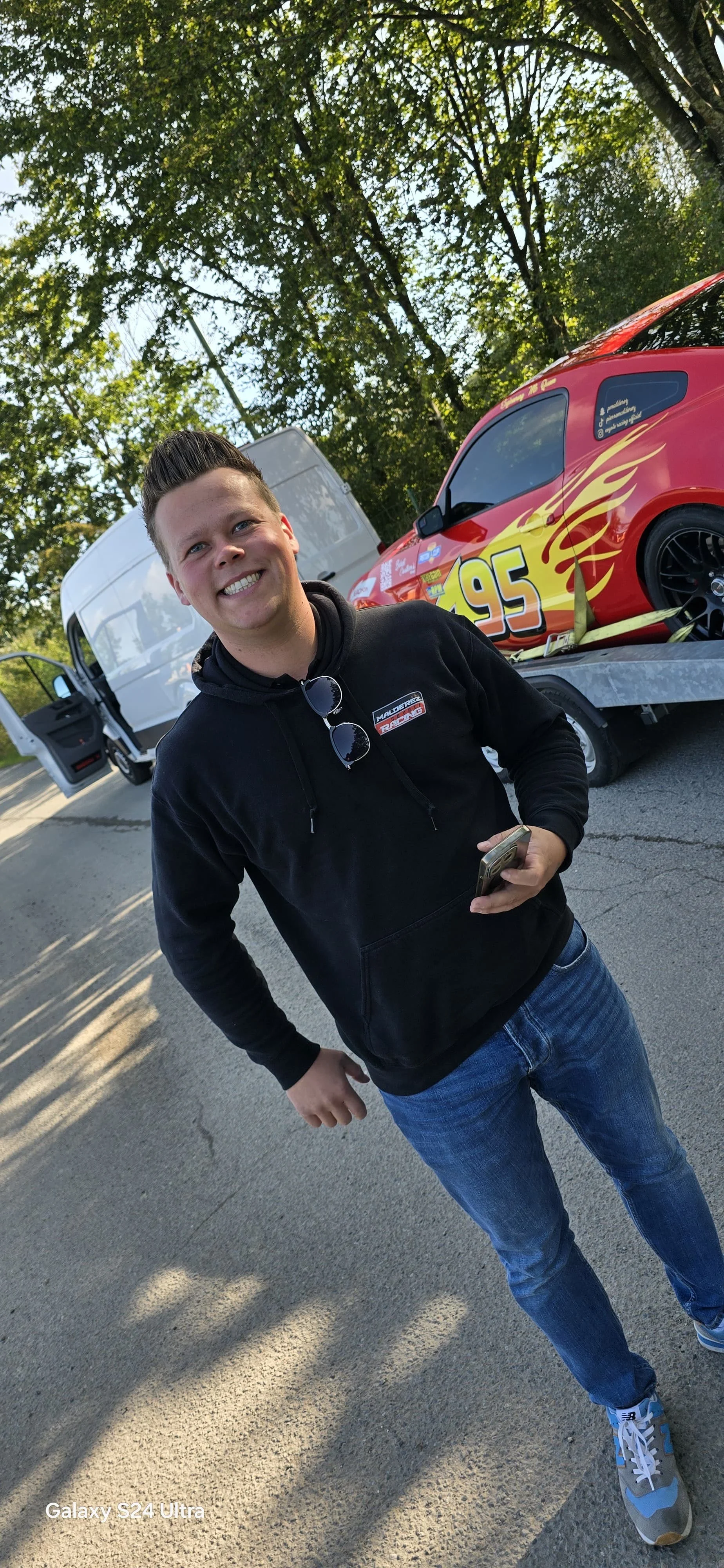Pierre Malderez souriant portant un sweat noir de sa production malderez srl , tenant un téléphone, en face d'une Ford Mustang de course rouge avec les numéros 95 et des flammes couleur de Flash Mc queen dans cars