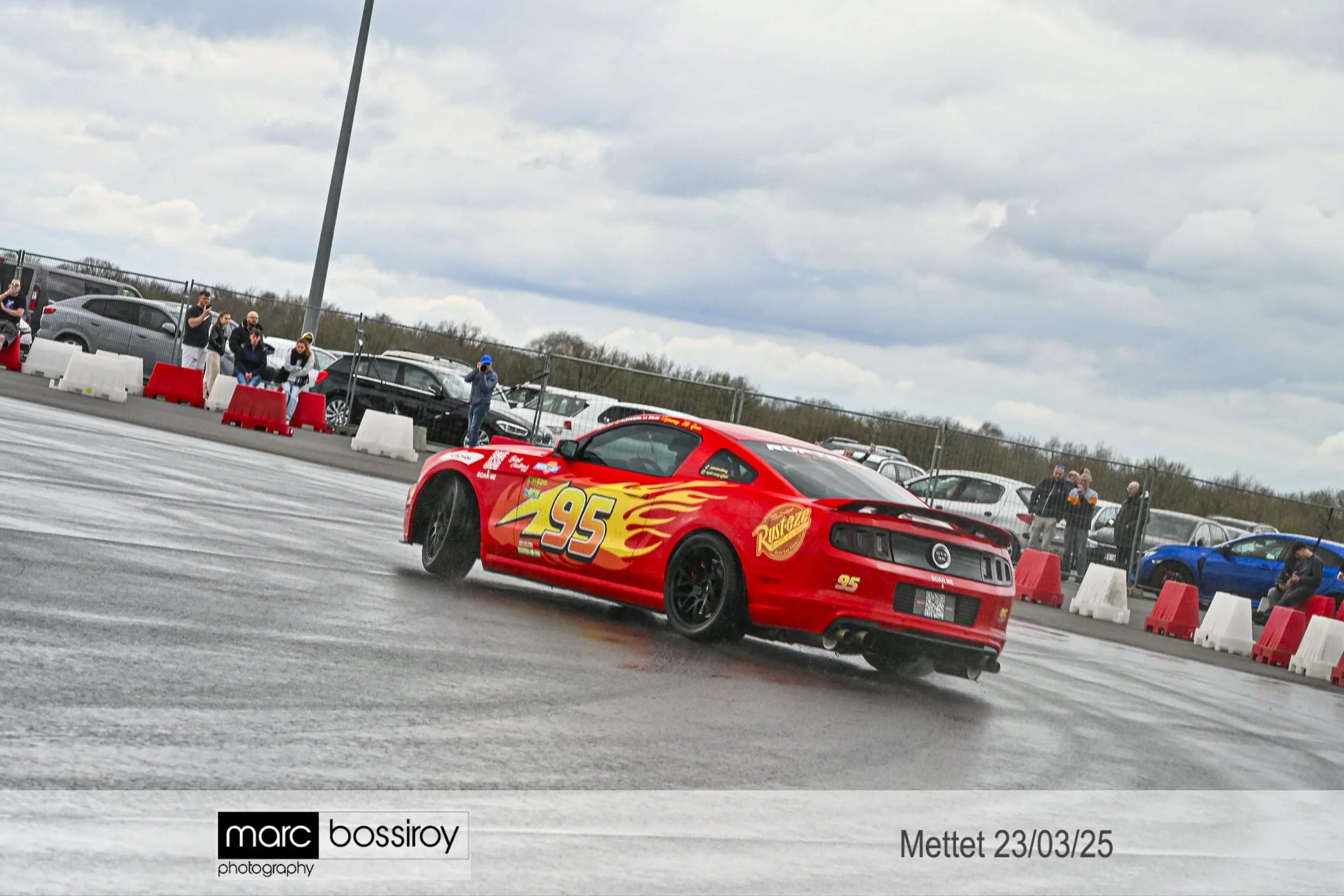 Ford Mustang de course rouge avec le numéro 95 et des flammes jaunes, en train de faire travers sur une piste de drift, avec des spectateurs en arrière-plan. Kachow