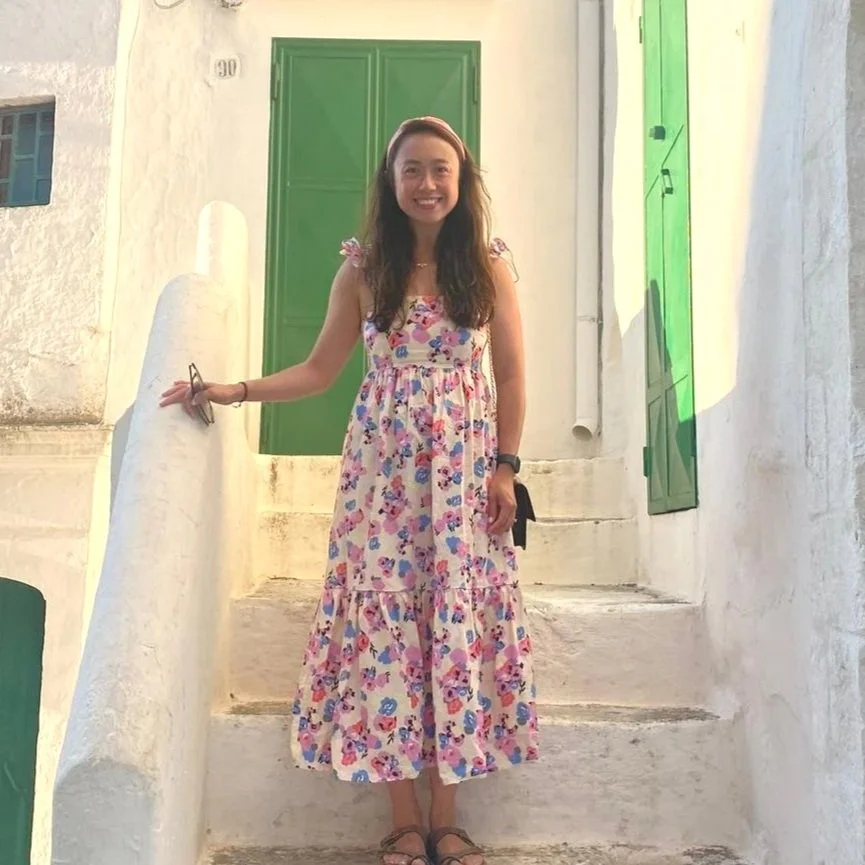 A woman with sunglasses and a pink sweater smiling next to a happy golden retriever dog outdoors, with green foliage in the background.
