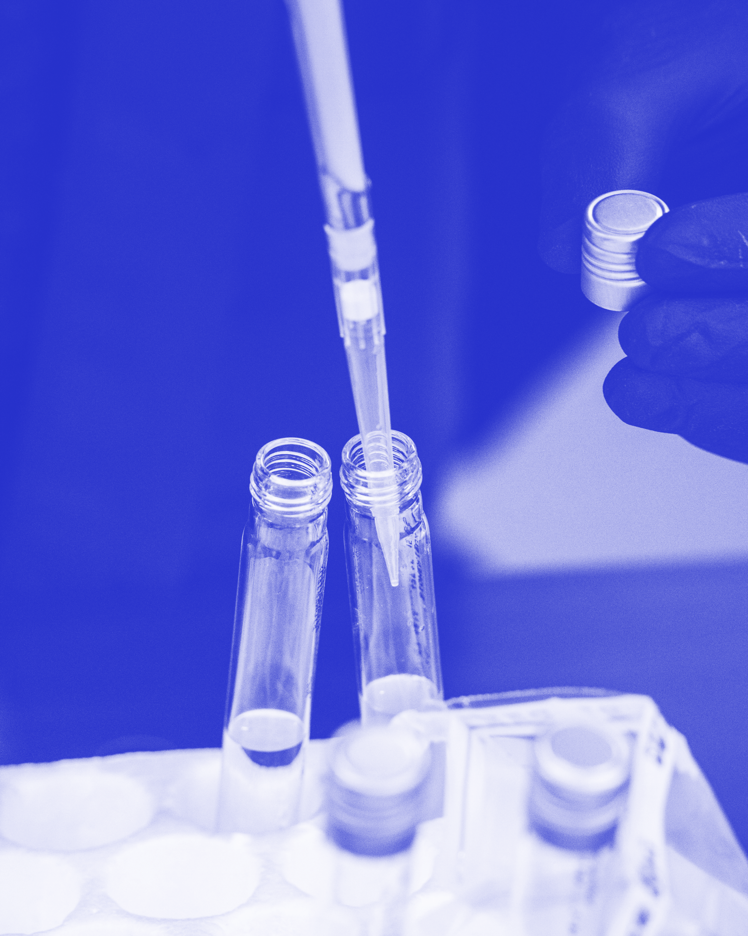 Laborant*in hält einen Schraubverschluss eines Reagenzglases in der einen Hand und eine Pipette, die eine Flüssigkeit in ein Reagenzglas gibt, in der anderen.