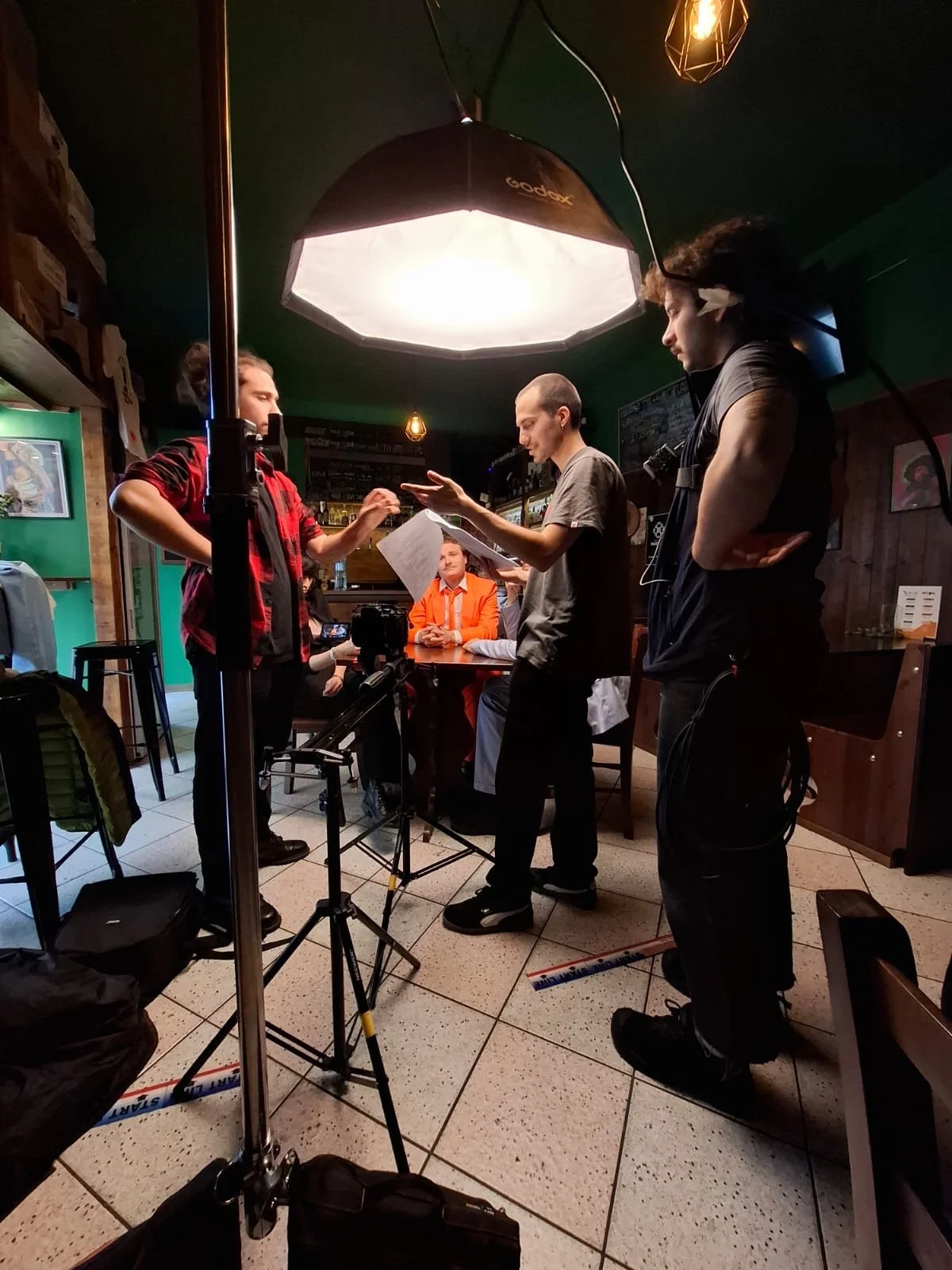 Un set cinematografico in un ristorante affollato, con alcune persone che discutono, una crew di produzione con attrezzature di illuminazione, microfoni e camere.