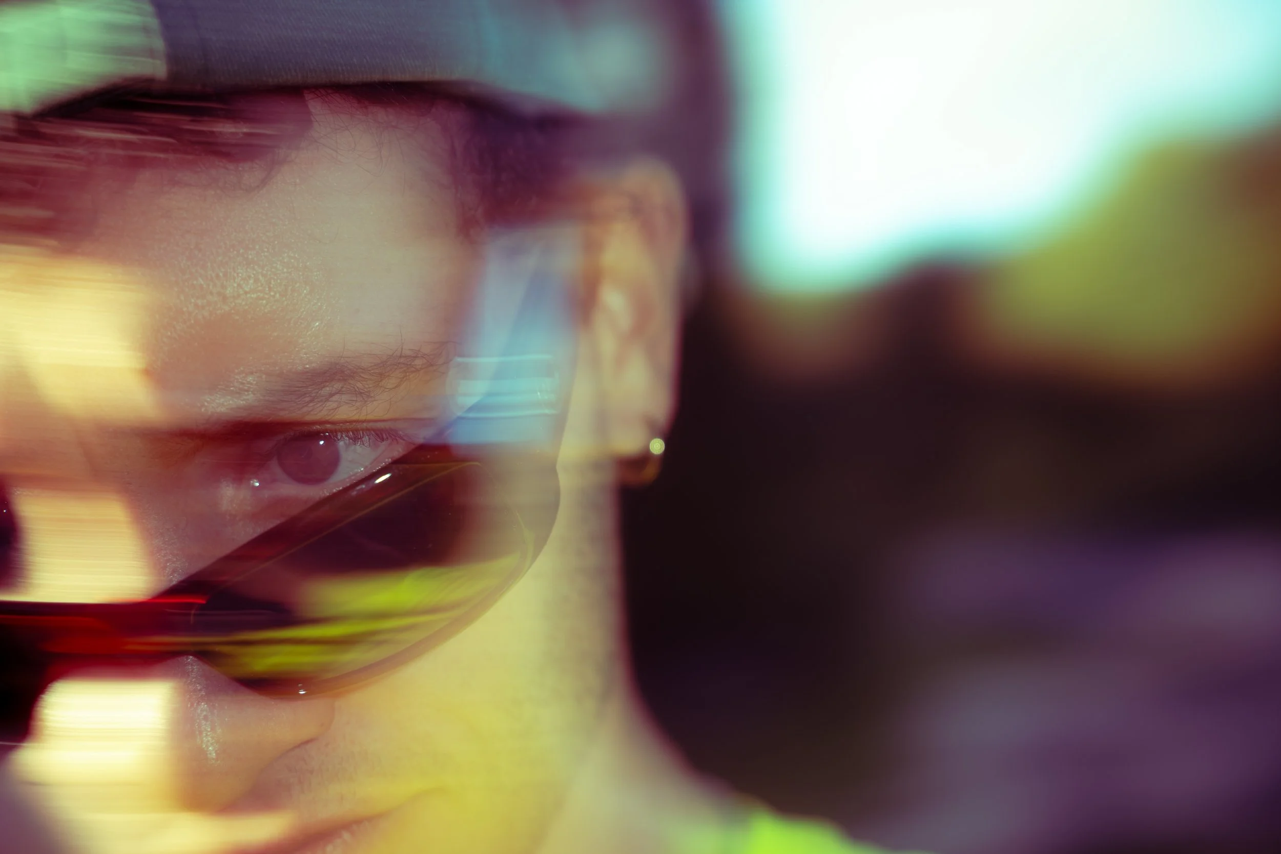 Ragazzo con occhiali da sole che guarda in basso, uno scatto con riflessi e sfocature di luci colorate.