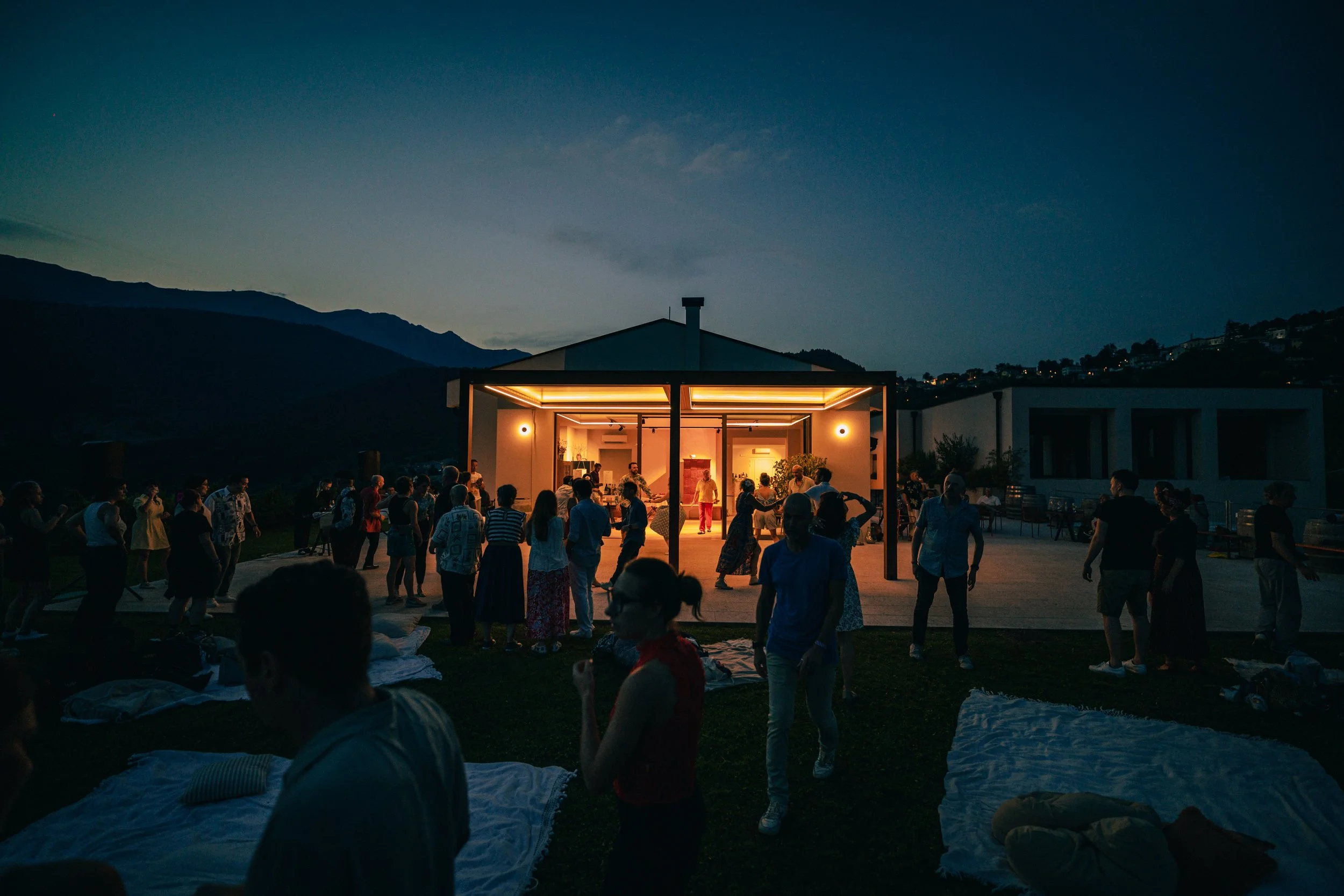 Festeggiamento serale con molte persone che ballano e socializzano all'aperto davanti a una casa moderna illuminata, con montagne sullo sfondo e un cielo al tramonto.