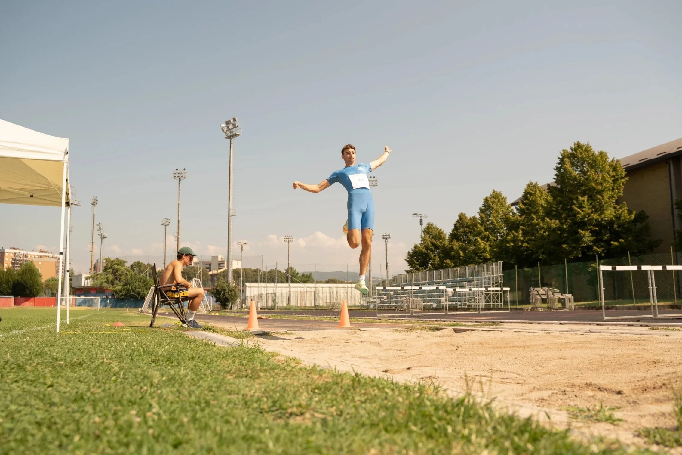 Un atleta sta saltando in lungo durante una gara all'aperto, con un pubblico e strutture sportive sullo sfondo.