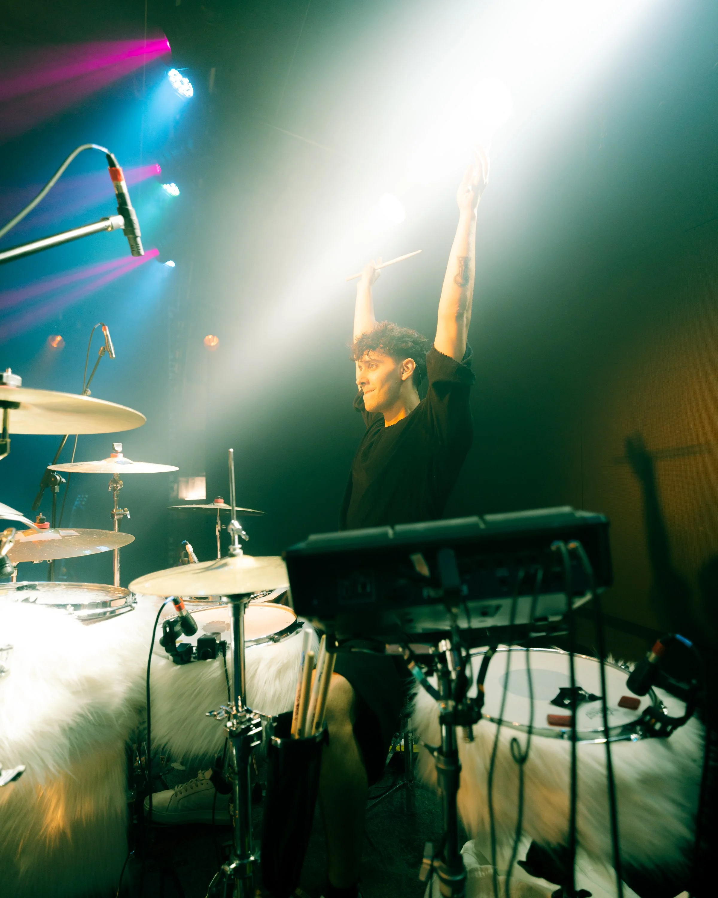 Un musicista dal vivo che suona la batteria mentre incita il pubblico, con luci colorate sul palco.