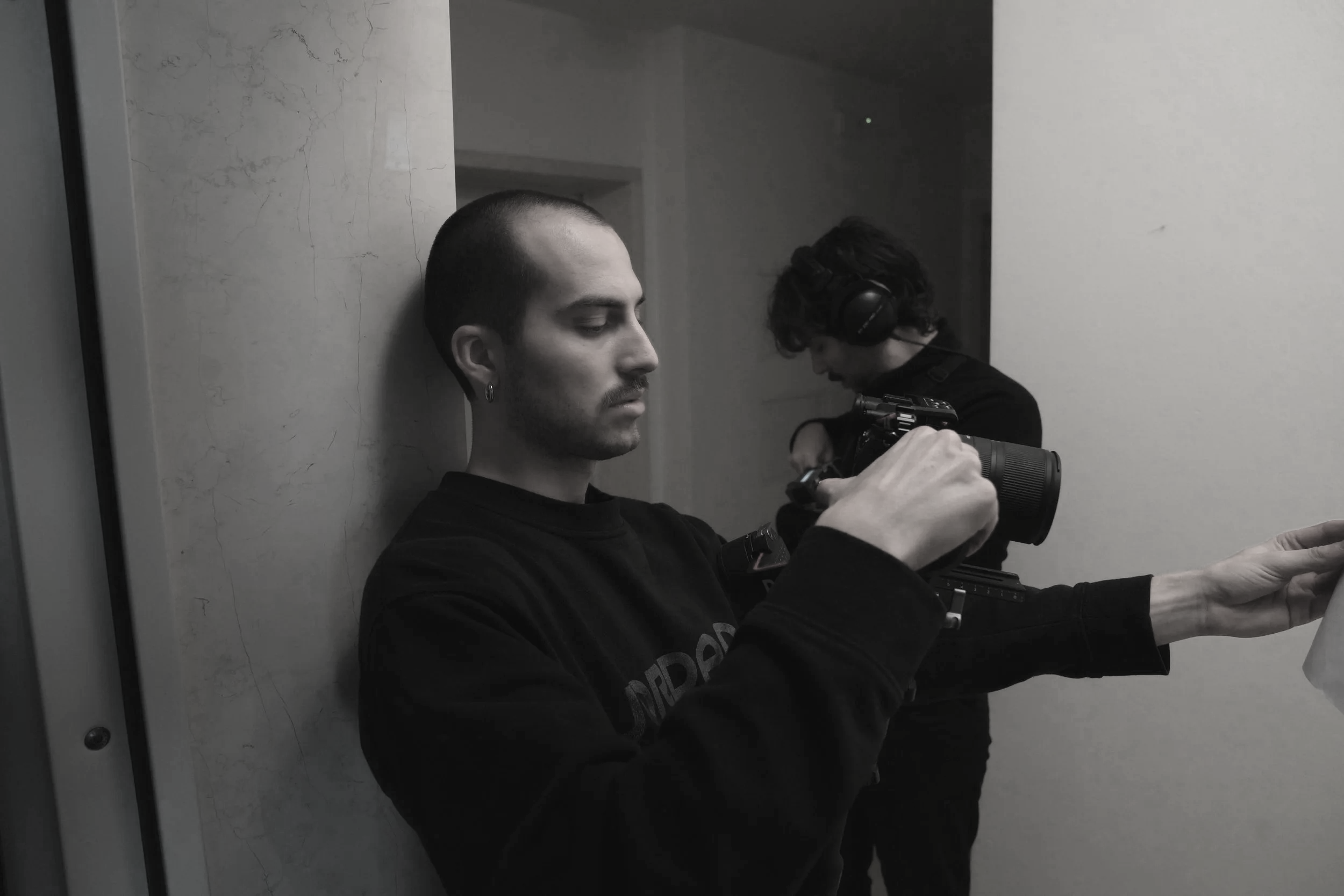 Due uomini in un ambiente interno, uno con capelli rasati e un orecchino, che guarda la ripresa della camera, e un altro con cuffie che controlla l'audio registrato.