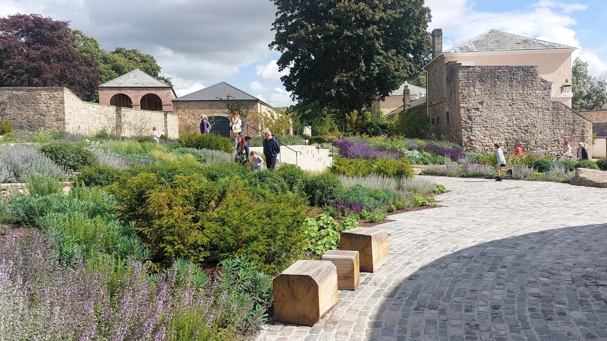 People walking along a cobblestone path in a lush garden with various plants and flowers, stone buildings and a large tree in the background, under a partly cloudy sky.