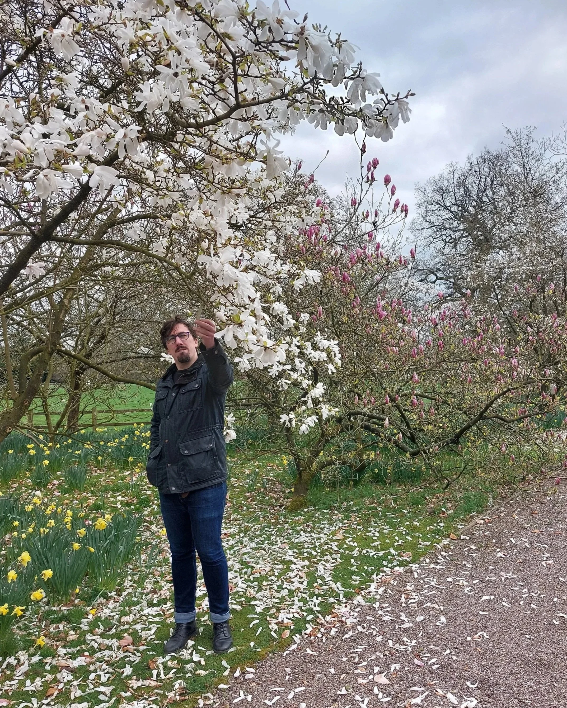 A man standing outdoors in a garden with blooming trees and yellow flowers, holding a branch with white flowers against a gray sky.