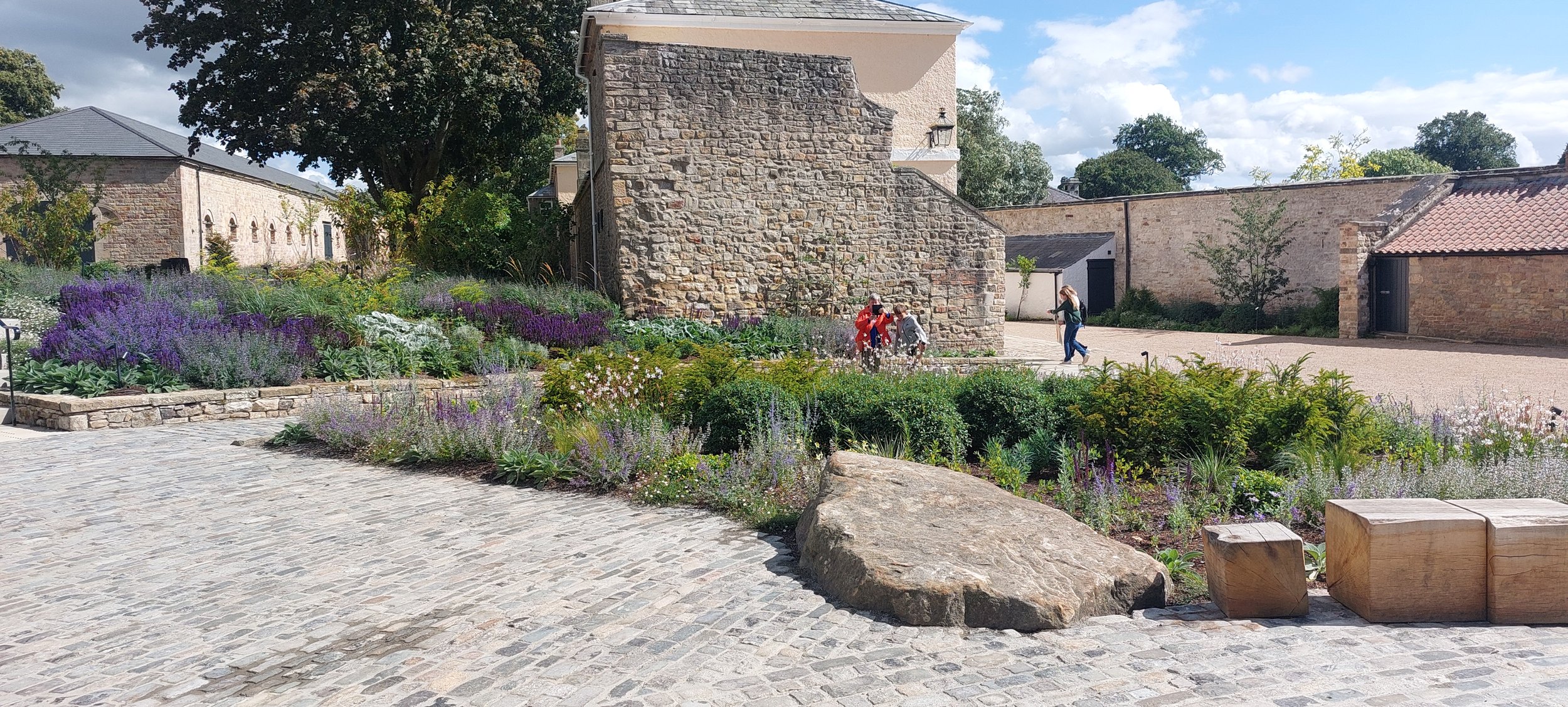 Cobblestone pathway in a garden with purple and green plants, large rock, and wooden bench in the foreground, stone walls and buildings in the background, and people walking in the distance.