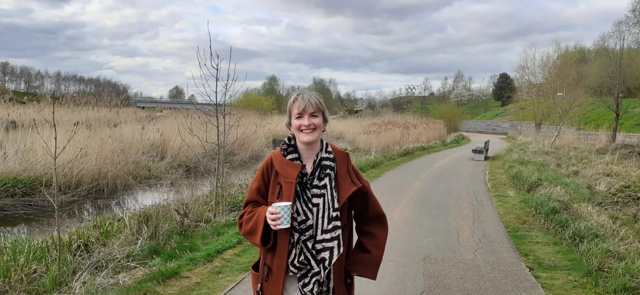 A smiling woman standing on a winding park pathway, holding a cup, with trees and grassy areas around her, and a cloudy sky overhead.