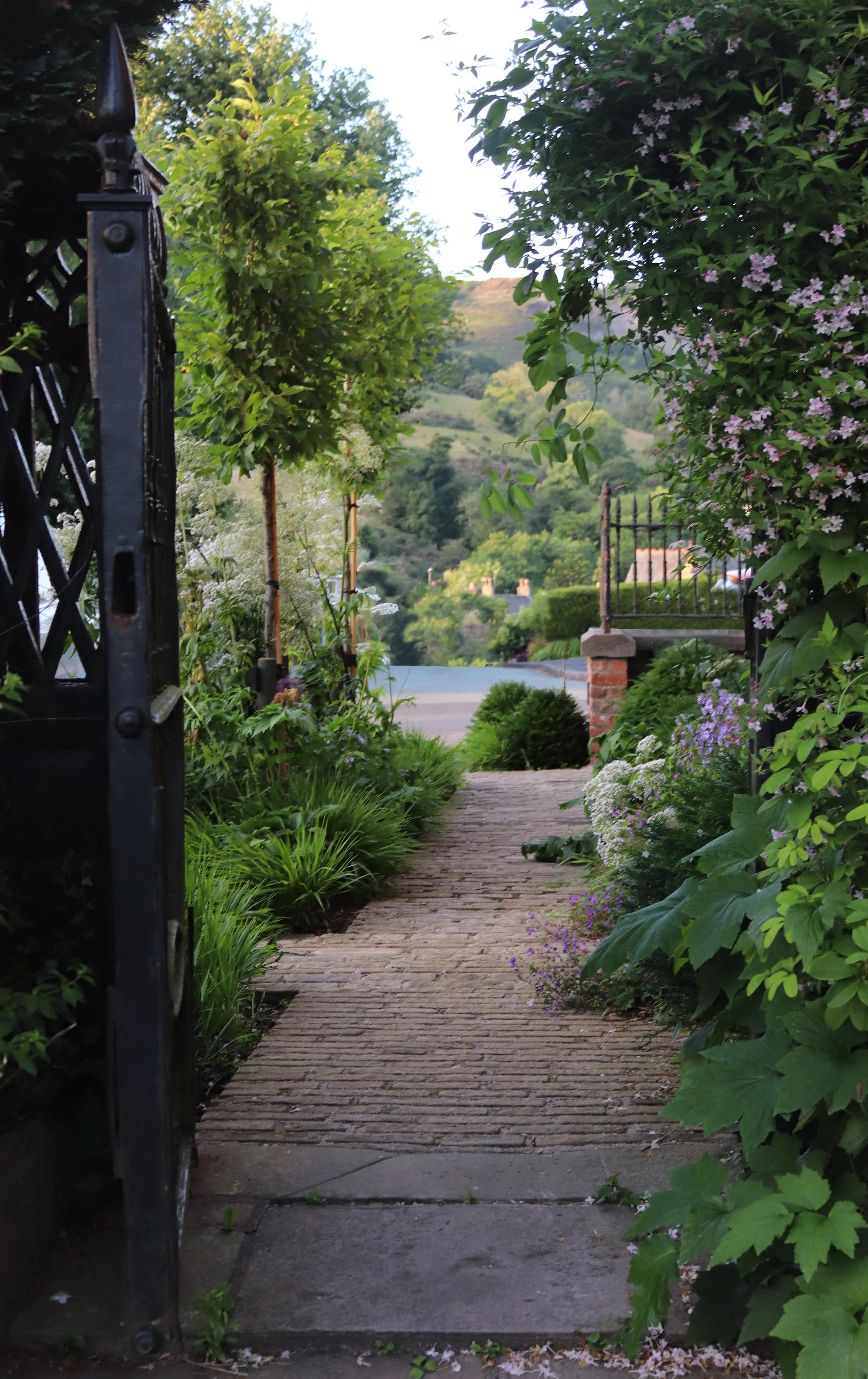 A brick pathway leading out from a garden gate, surrounded by lush flowering plants and greenery, with mountains and rooftops in the background.