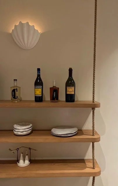 Wooden floating shelves with four bottles of wine, stacked white plates, and a small decorative shoe on the bottom shelf. A white shell-shaped wall sconce above the shelves.