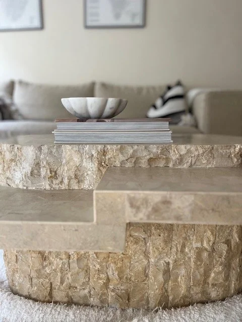 Close-up of a round beige stone coffee table with a bowl and magazines on top, in a living room with a beige sofa and framed pictures on the wall in the background.
