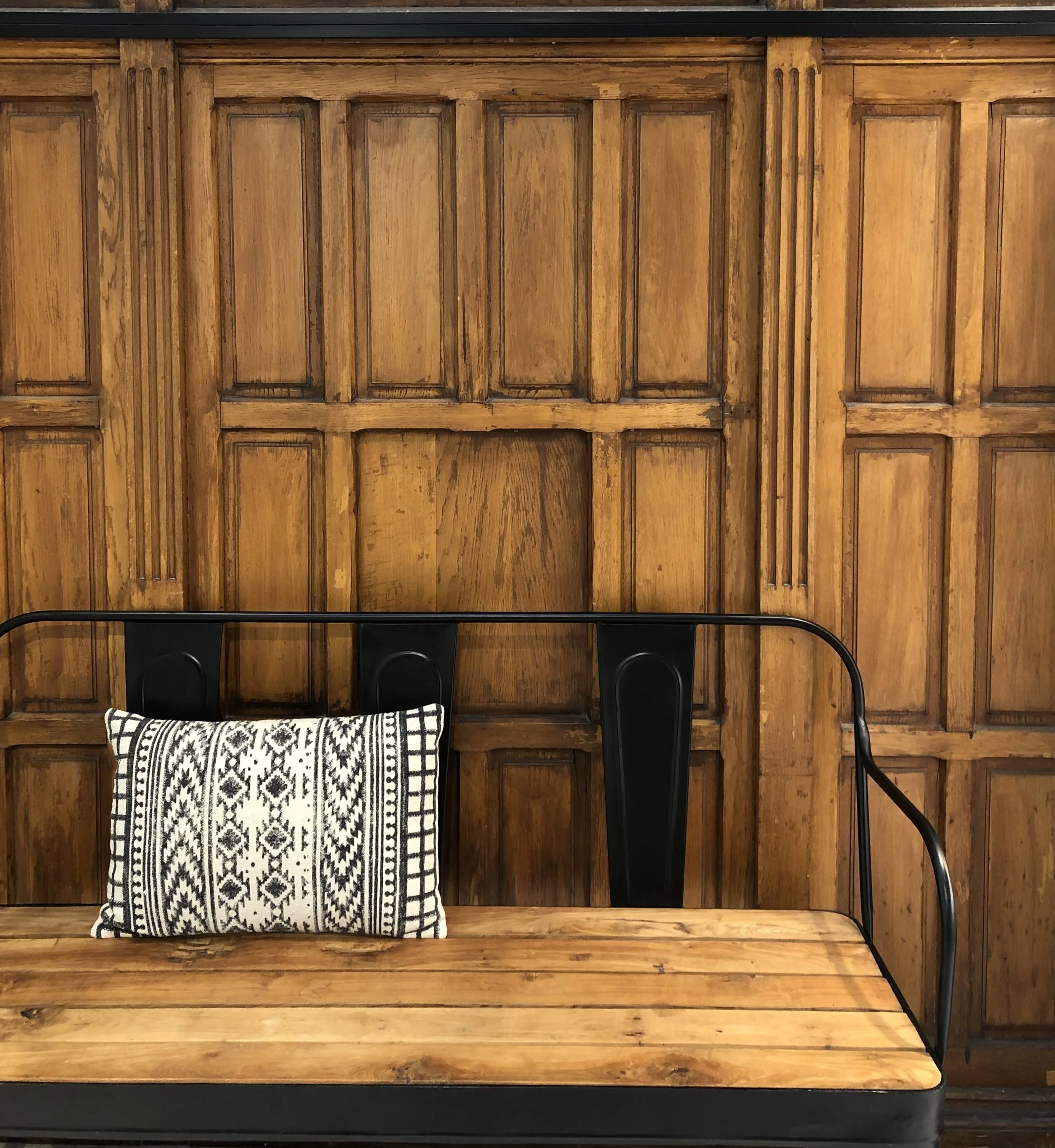 A wooden bench with a decorative black and white pillow, set against a wood-paneled wall with vertical and horizontal rectangular patterns.