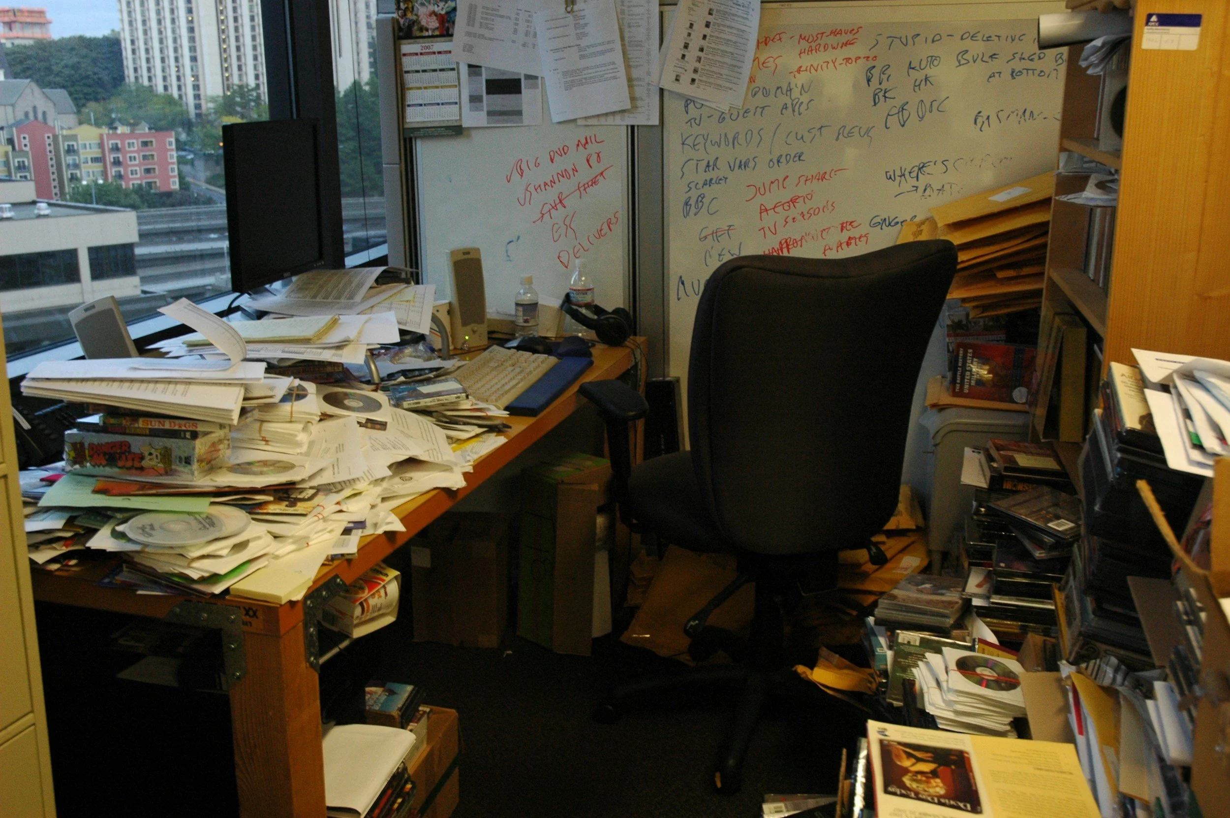 A cluttered office desk with piles of papers, CDs, and books, an office chair, a whiteboard with handwritten notes, and a window showing city buildings outside.