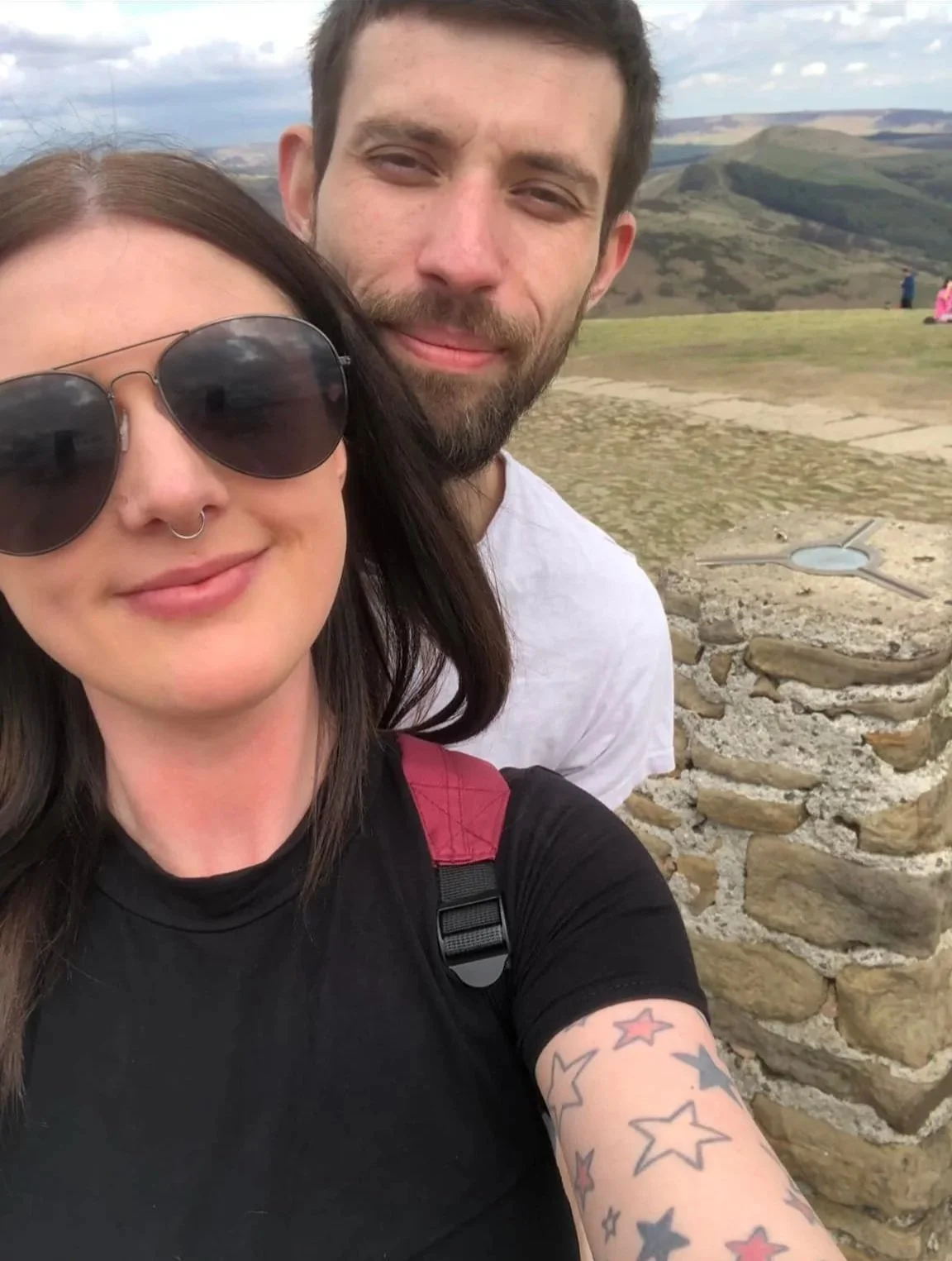 A woman and a man taking a selfie outdoors on a hilltop with rolling hills in the background. The woman has dark hair, sunglasses, a nose ring, and tattoos on her arm. The man has a beard and is smiling slightly.