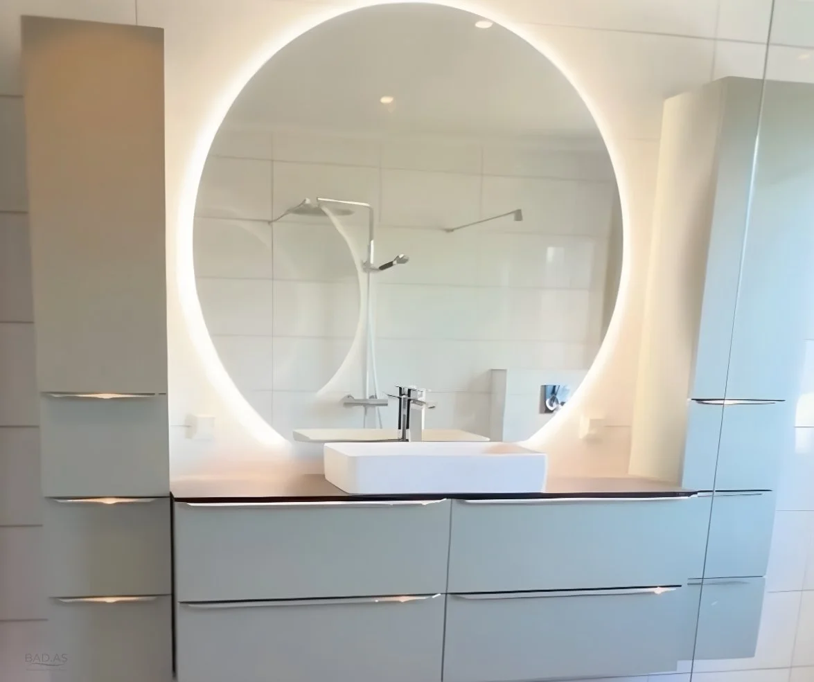 Et moderne bad med rund speil med innebygd belysning, hvit vask og speilskap med glatte fronter.