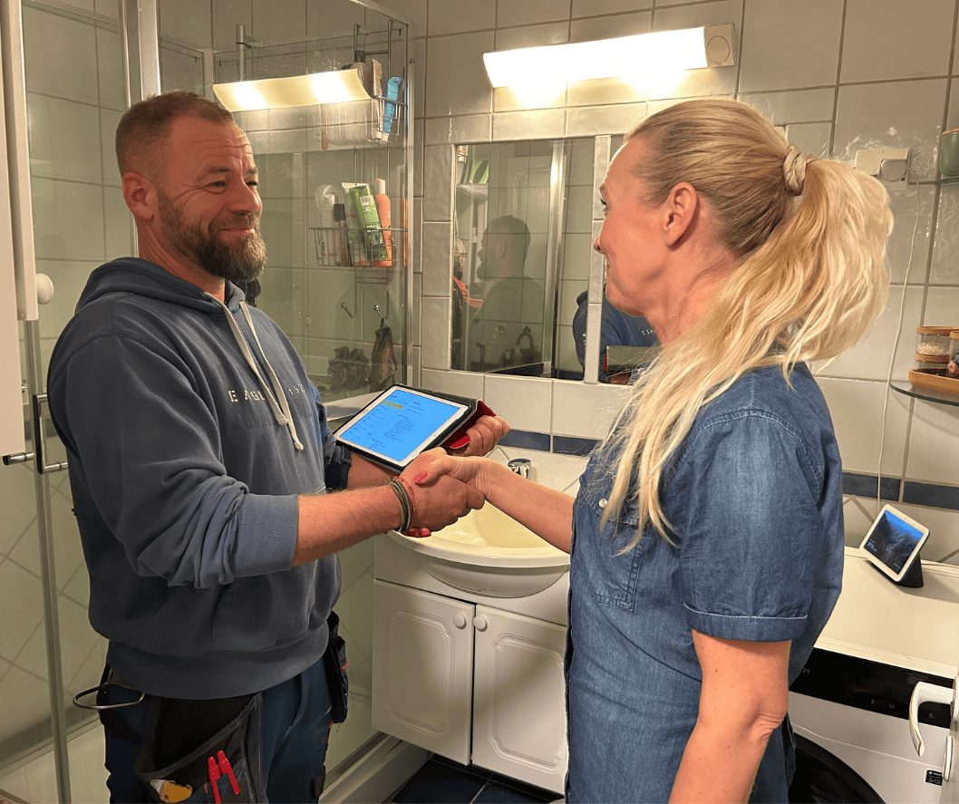 En mann og en kvinne utveksler håndtrykk i et baderom. Mannen holder en digital enhet, og kvinnen har langt blondt hår og bruker en blå skjorte. Bak dem er det speil og vask, og på hyller er det noen produkter.