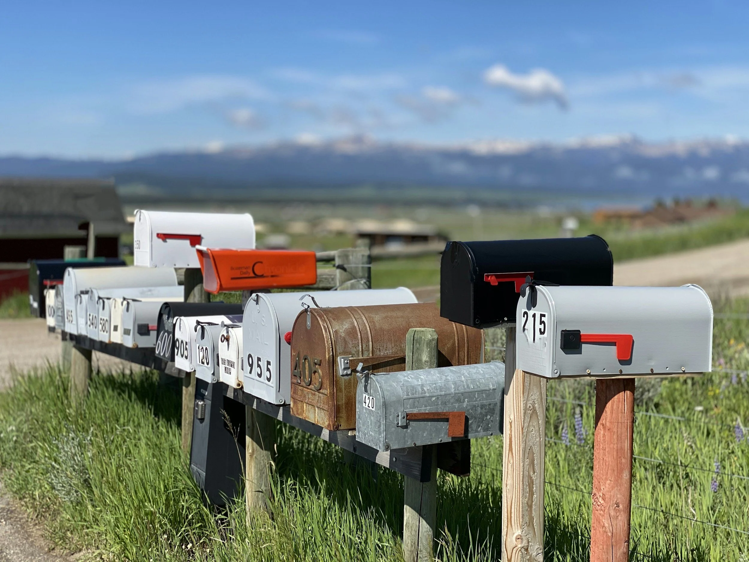 Multiple mailboxes of various colors and shapes mounted on wooden posts along a rural roadside with green grass. The background includes a scenic landscape with distant mountains, a partly cloudy blue sky, and a few scattered buildings.