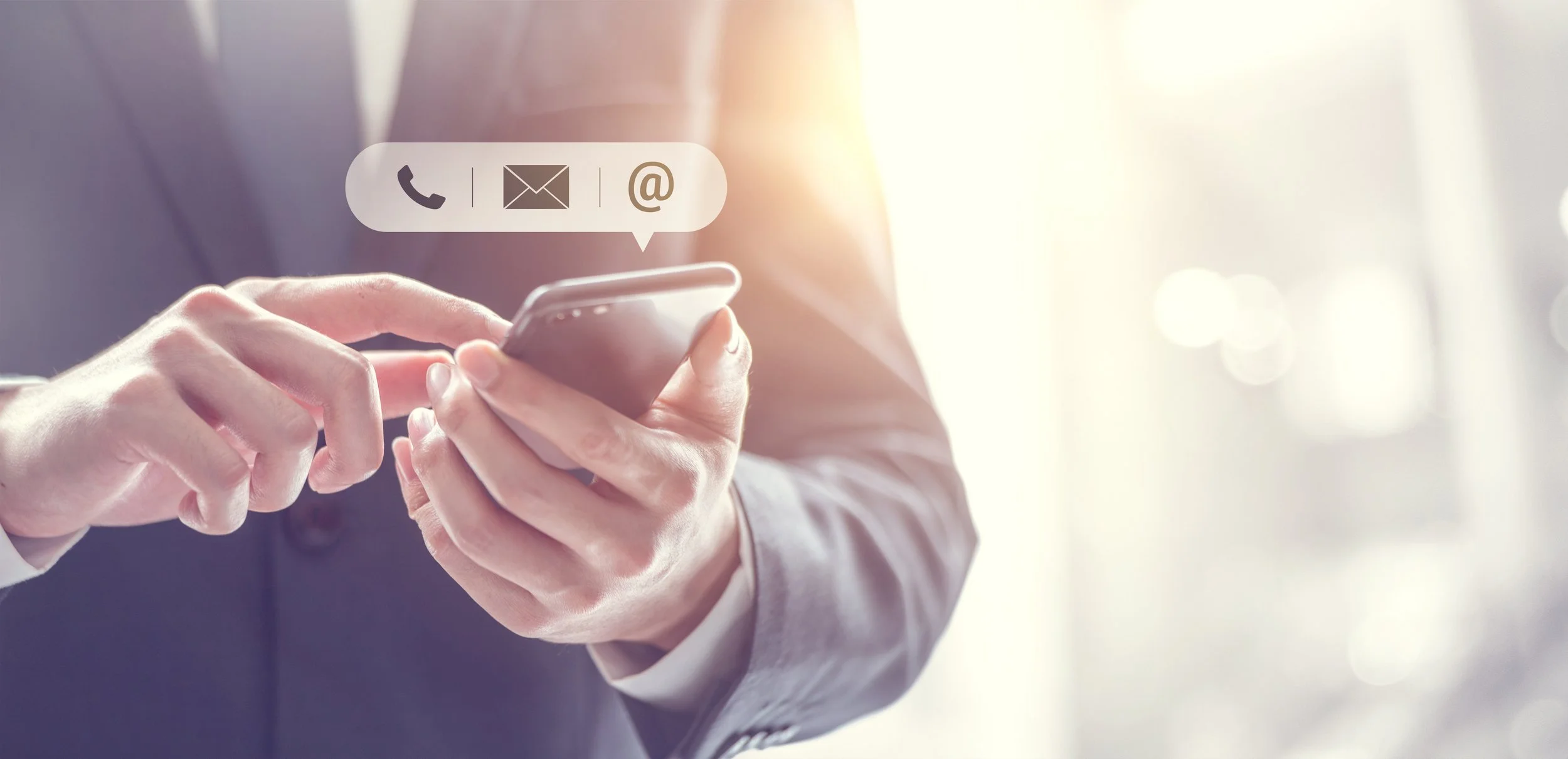Person in business attire using a smartphone with communication icons—phone, email, and at symbol—above their hand.