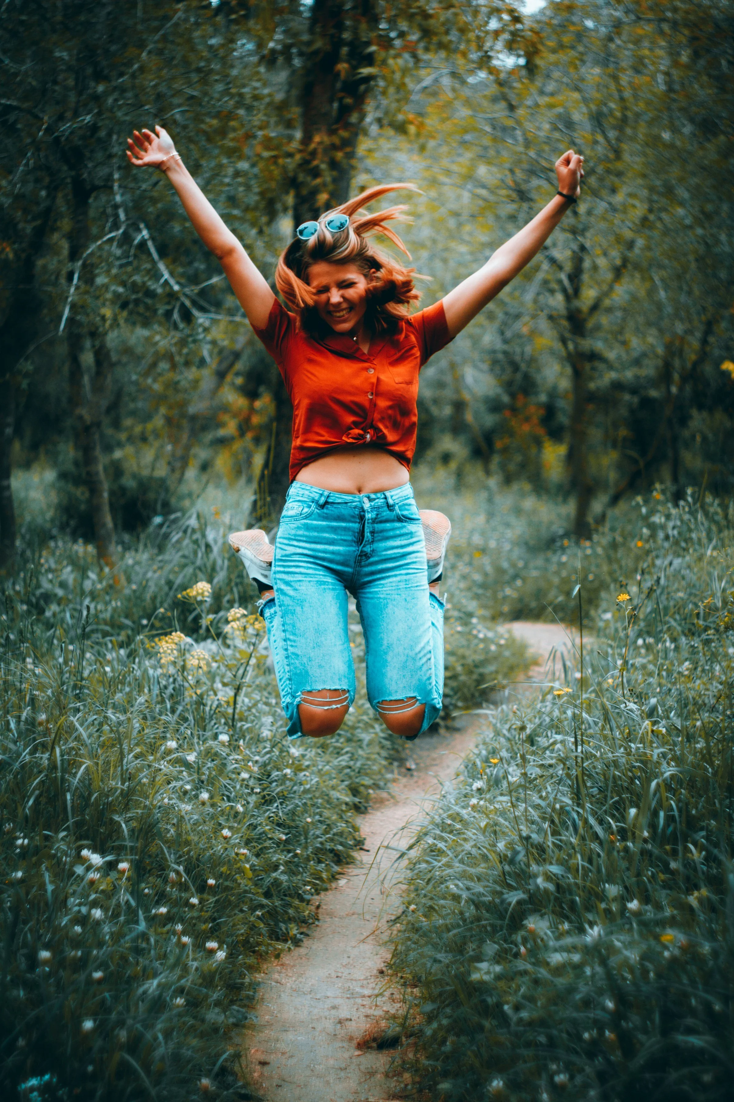 A young woman with red hair jumping in the air, with arms raised and smiling, on a narrow dirt trail surrounded by green grass and wildflowers in a forest.