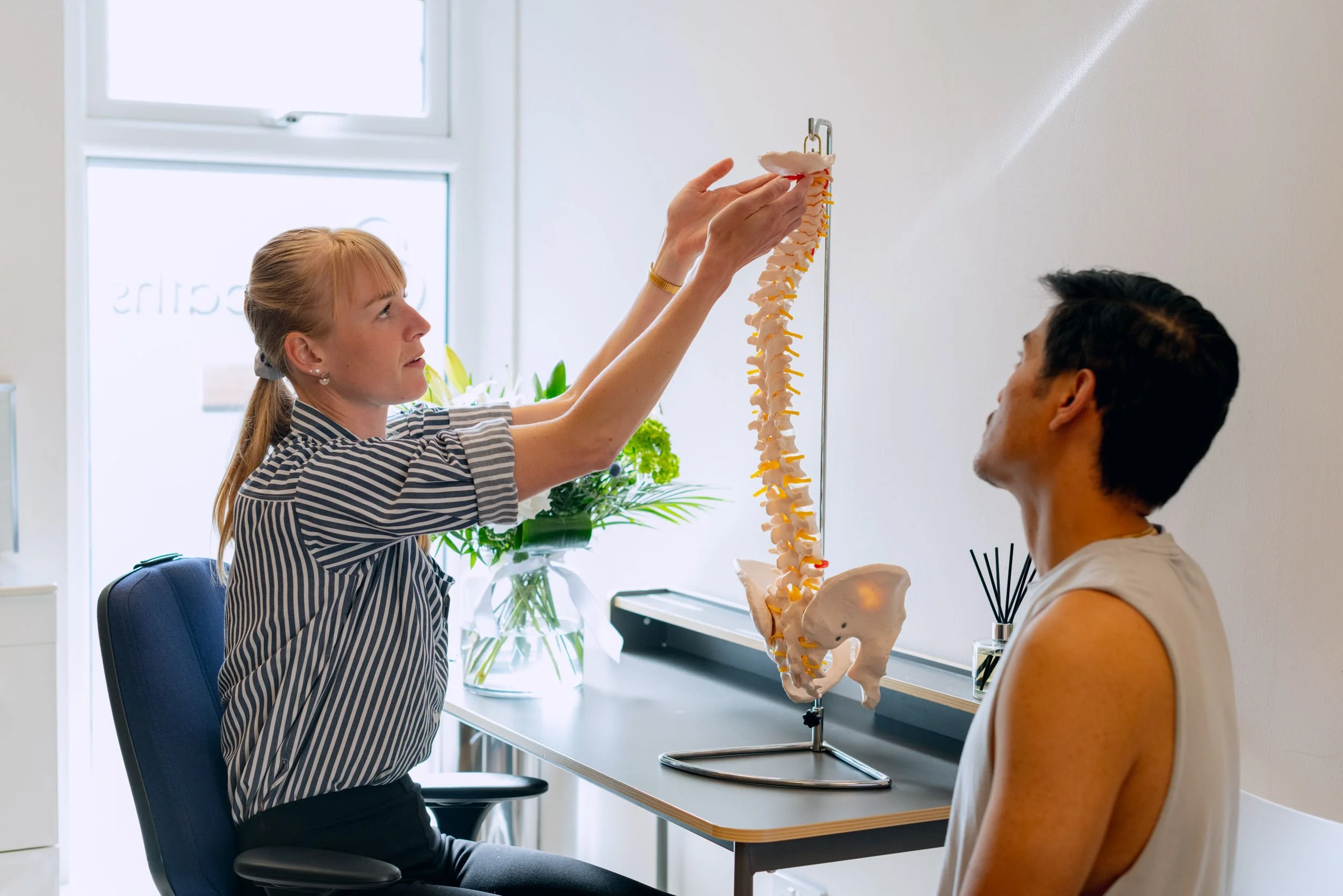 A woman showing a model of a human spine to a patient in a medical office, with a window and plants in the background.