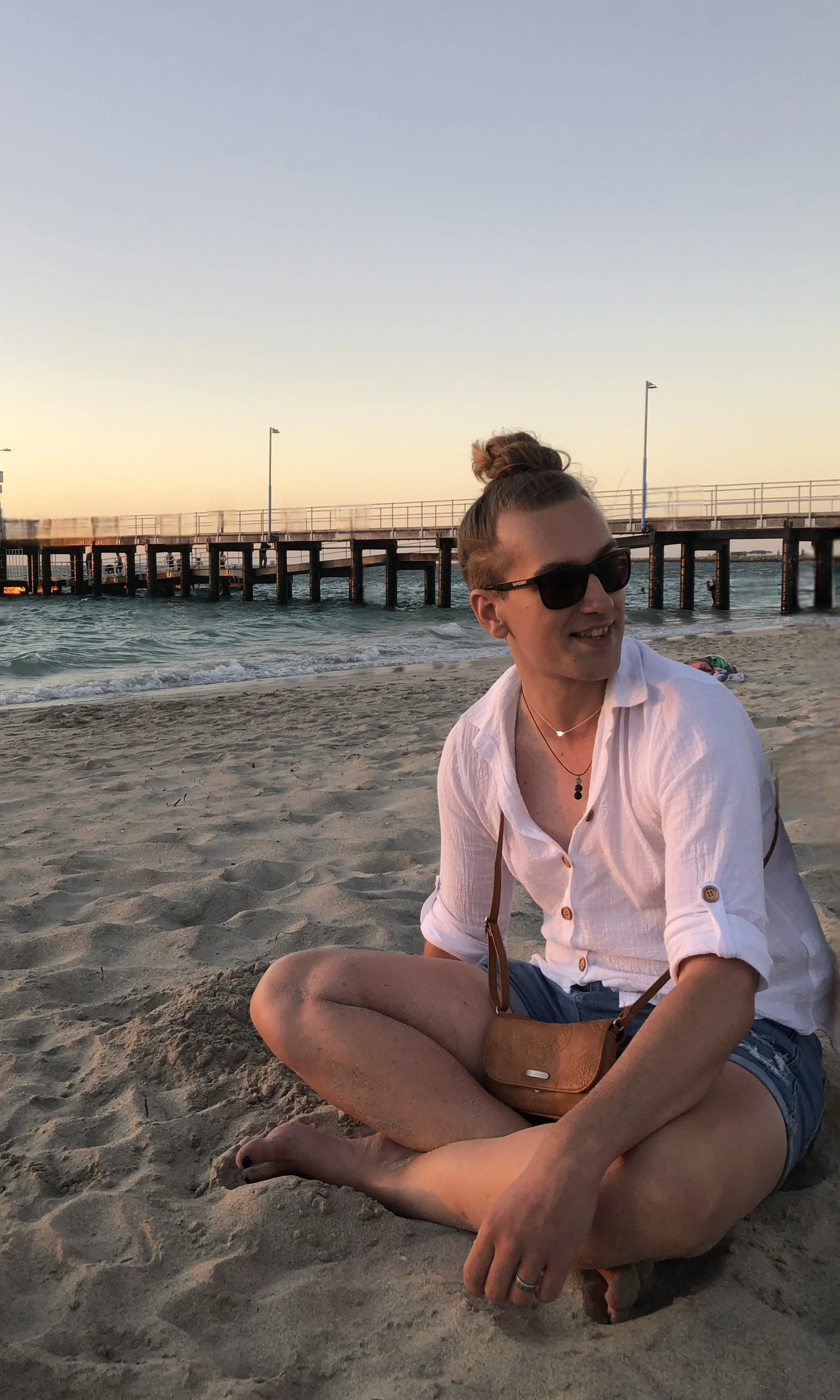 A woman sitting on the beach with a pier in the background during sunset. She is smiling, wearing sunglasses, a white shirt, and denim shorts, with a brown purse across her shoulder.