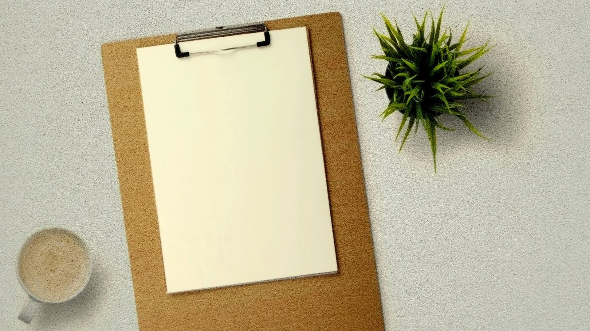 Top-down view of a workspace with a clipboard holding blank paper, a potted plant, and a cup of coffee on a light-colored surface.