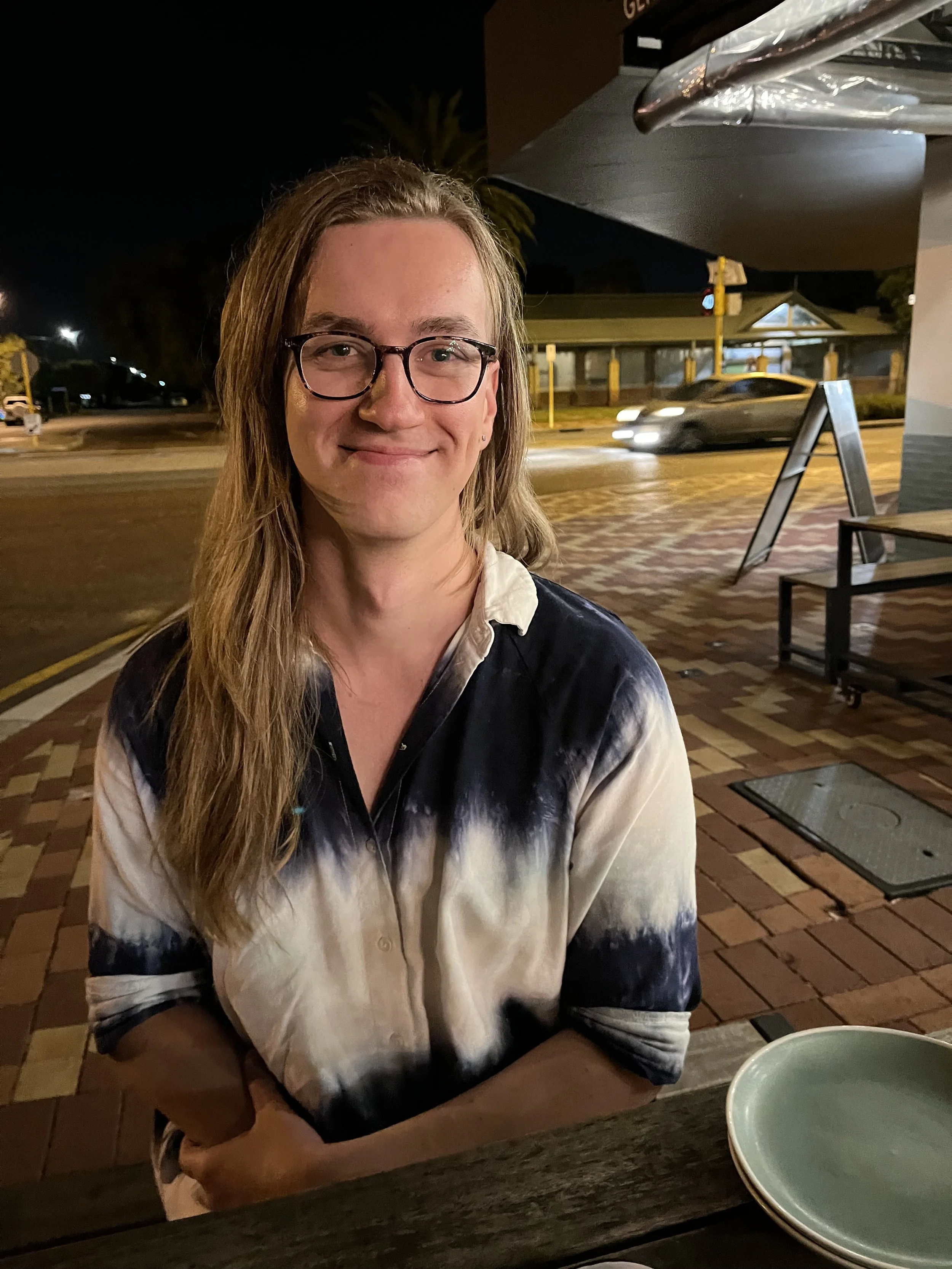 A woman with long, light brown hair, glasses, and a tie-dye shirt sitting at an outdoor table at night with a street and cars in the background.