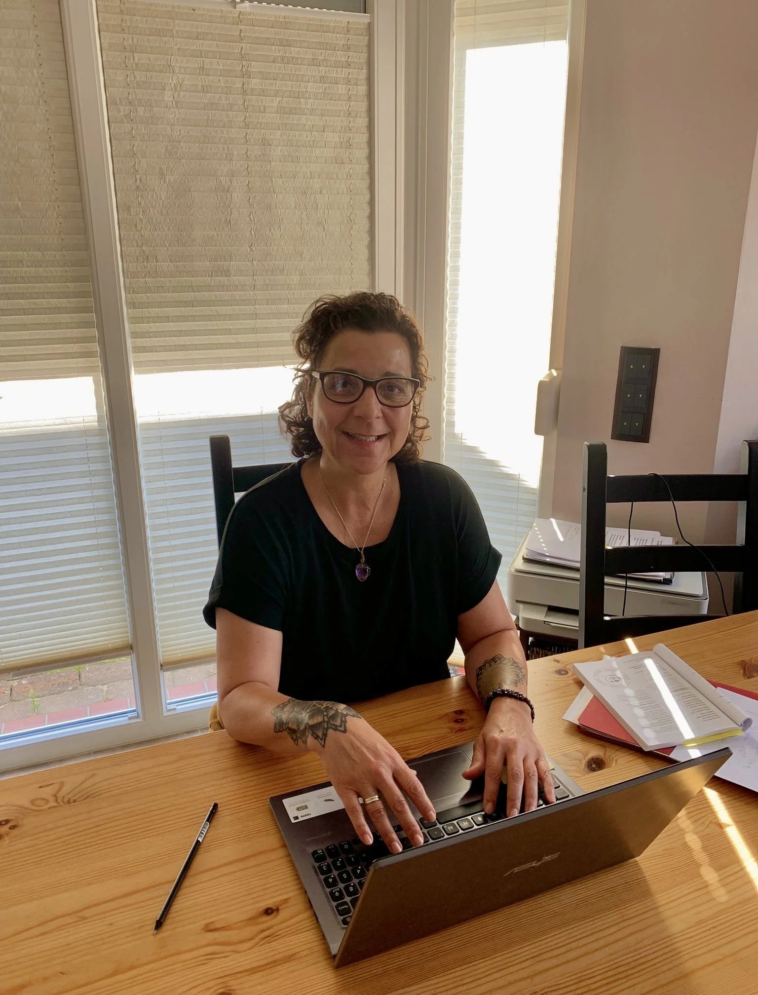 Frau mit Tattoos auf Armen, Brille, schwarz Kleid, sitzt an Holztisch mit Laptop, Papier und Stift, hinter große Fenster mit Rollos in hellen Tönen.