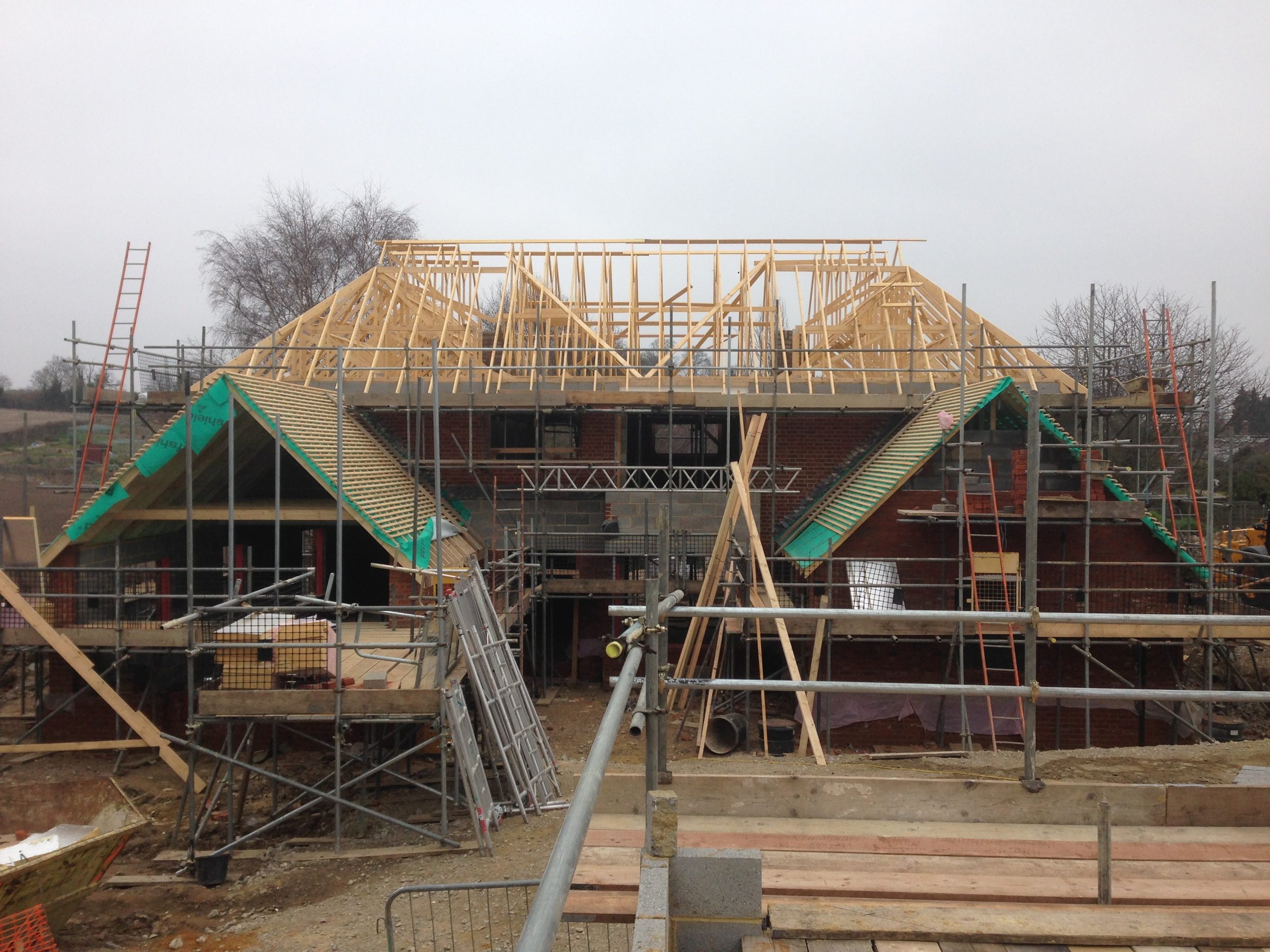 Construction site of a house with scaffolding and wooden roof framing in progress, brick walls visible, overcast sky.