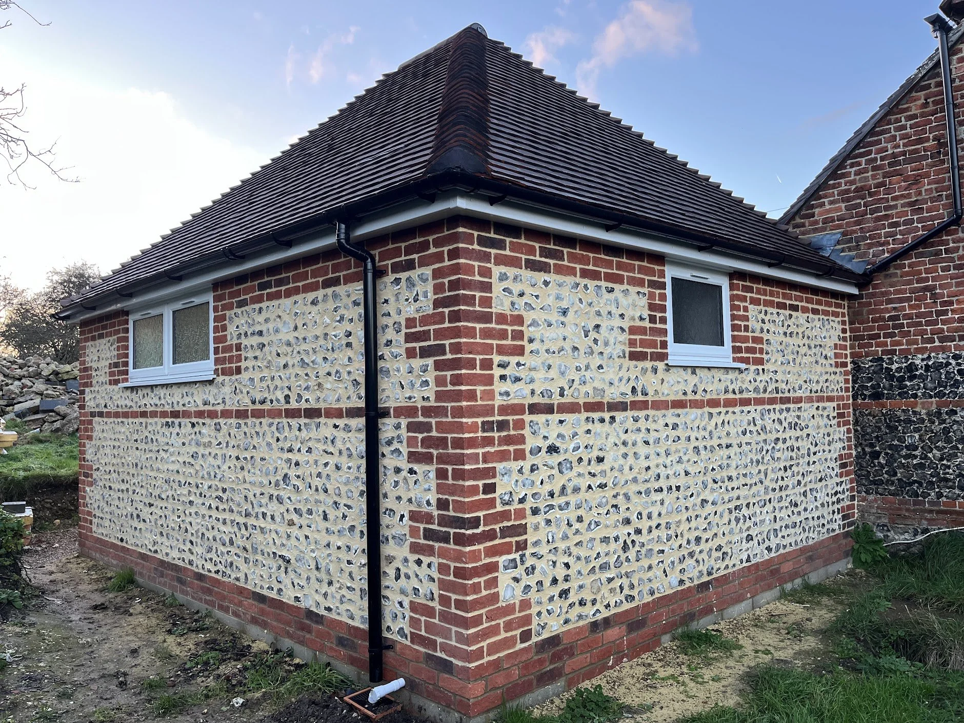 A brick house with a pebble and brick exterior wall, two small windows, and a brown tiled roof with black gutters, flint walls in Alkham Valley, Kent. 
