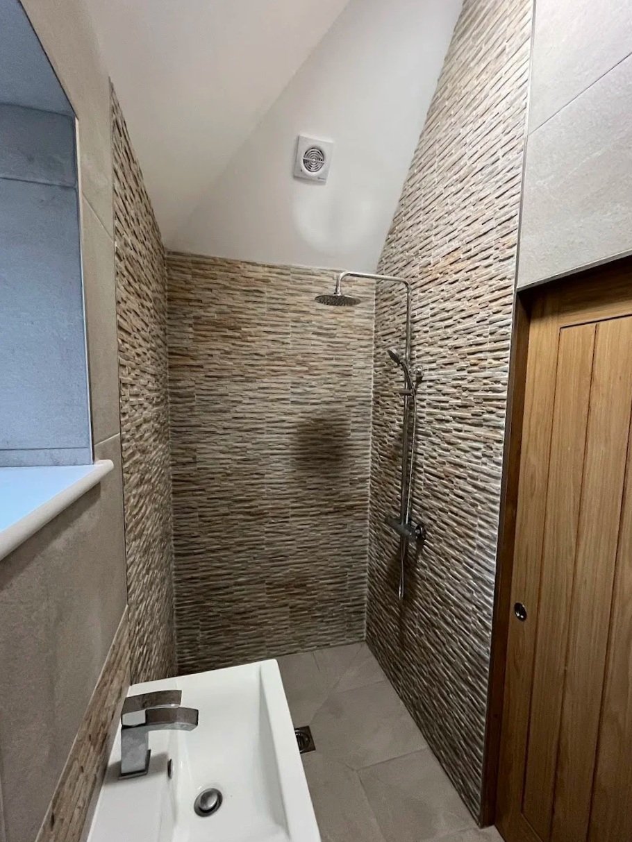 A small bathroom with a walk-in shower featuring textured brown tiles, a wooden door, and a small white sink with a modern faucet, new bathroom in Alkham. 