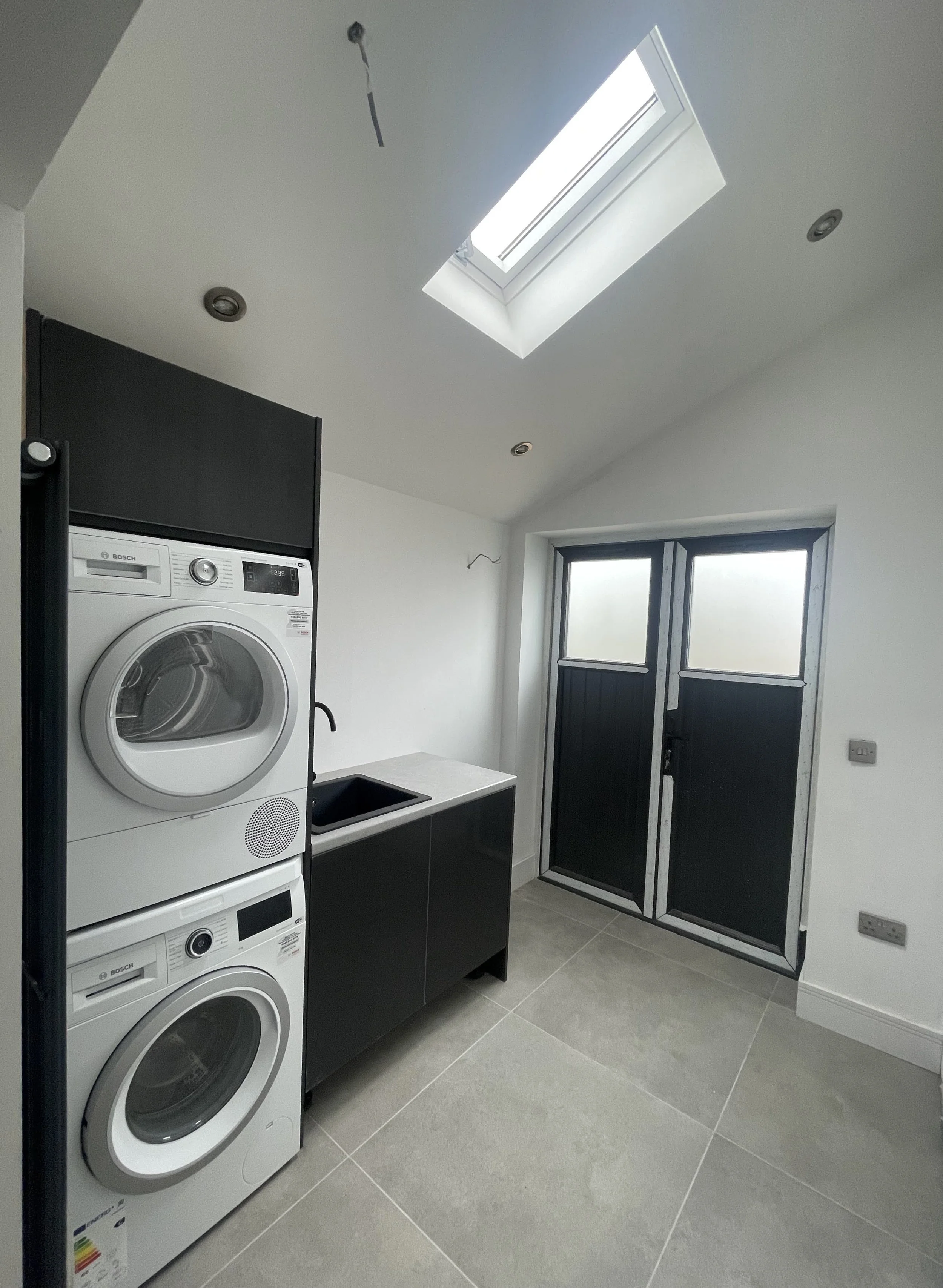 A laundry room with stacked washing machines and dryer, a black sink, a windowed door, a skylight, and white walls.