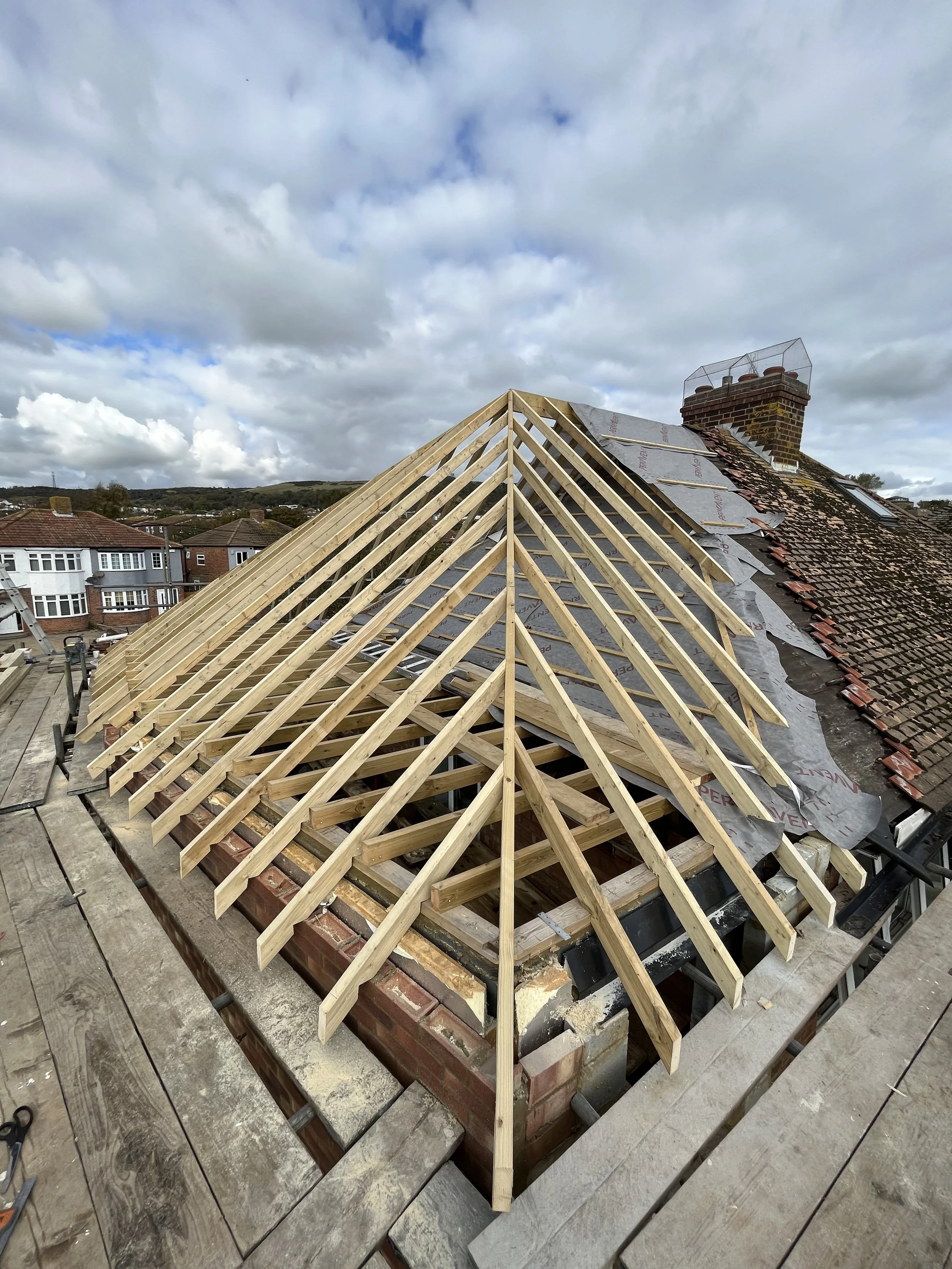 Roof under construction with new wooden trusses installed, part of the old tiled roof still visible.