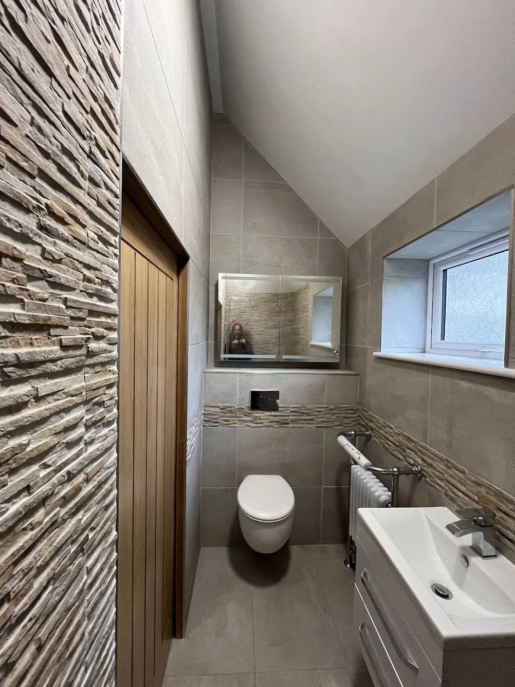 A small modern bathroom with beige tiled walls and a sloped ceiling. There is a white toilet, a small white sink with a cabinet, a mirror above the toilet, a window on the right, and a textured stone accent wall on the left, new bathroom in Alkham, K