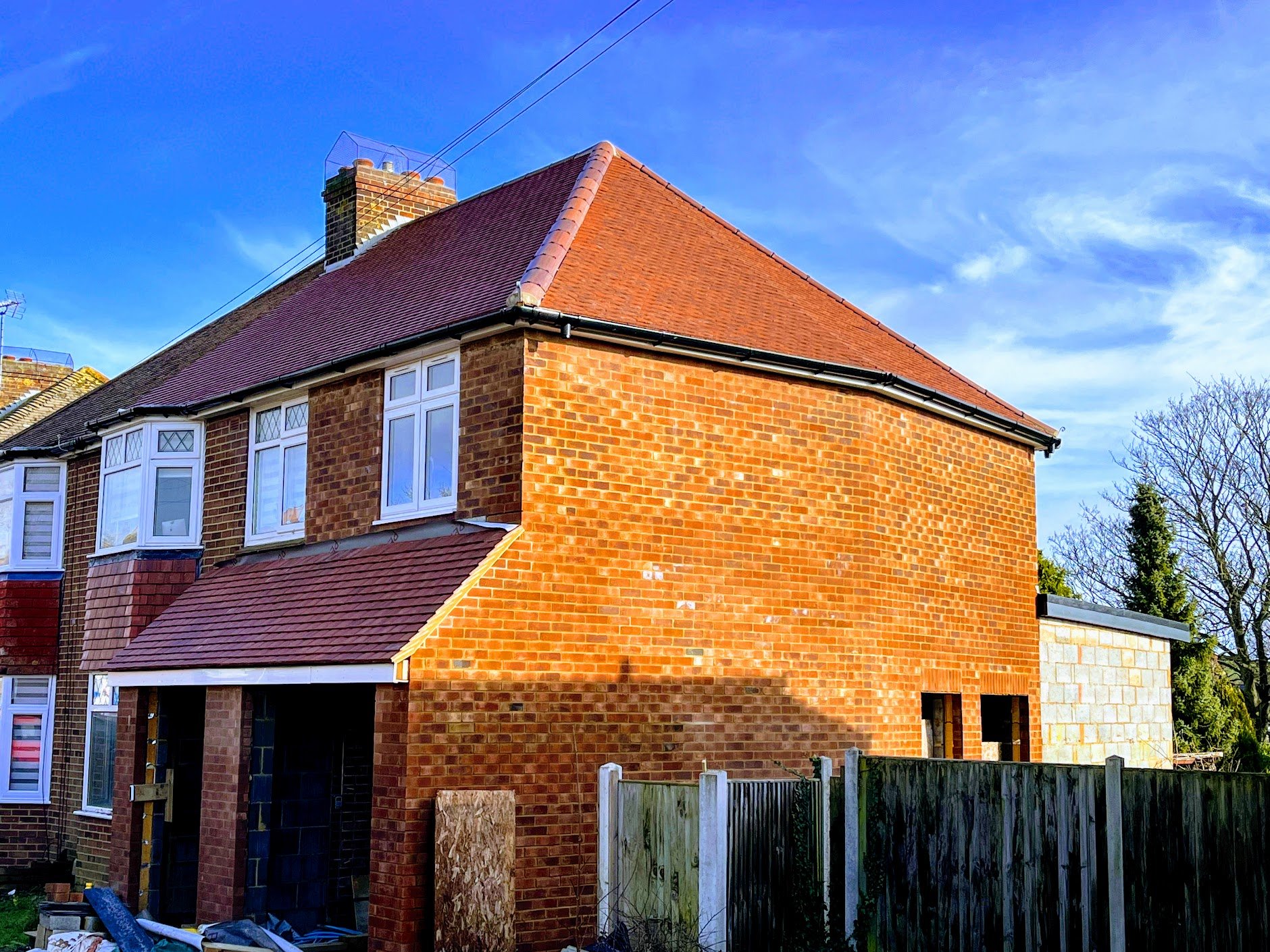 Side view of a two-story red brick house with a pitched red tile roof, partially under construction, beside a wooden fence, with blue sky and trees in the background, New Roof in Folkestone, Kent. 