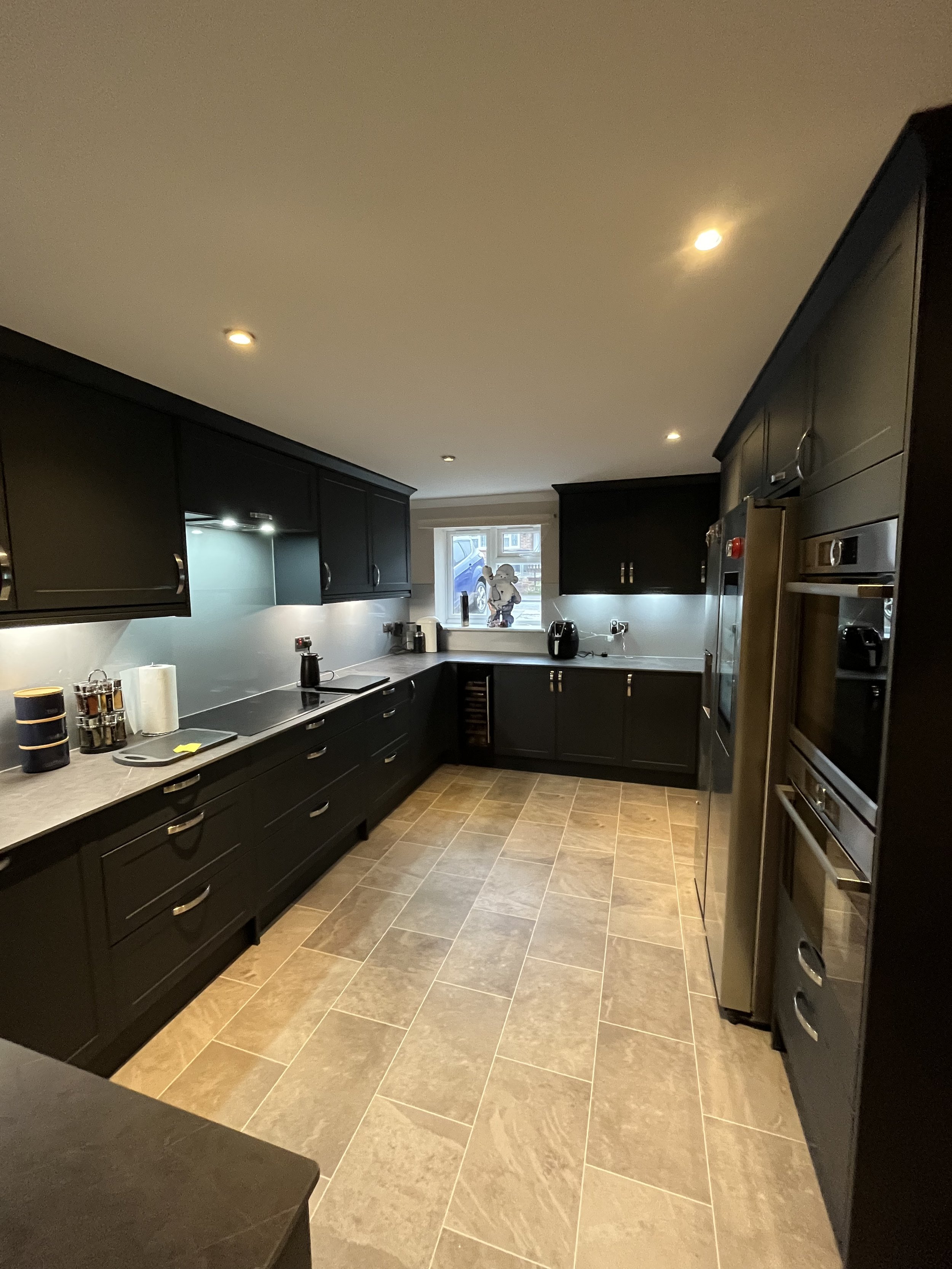Modern black kitchen with beige tile flooring, stainless steel appliances, and under-cabinet lighting, viewed from the entrance.