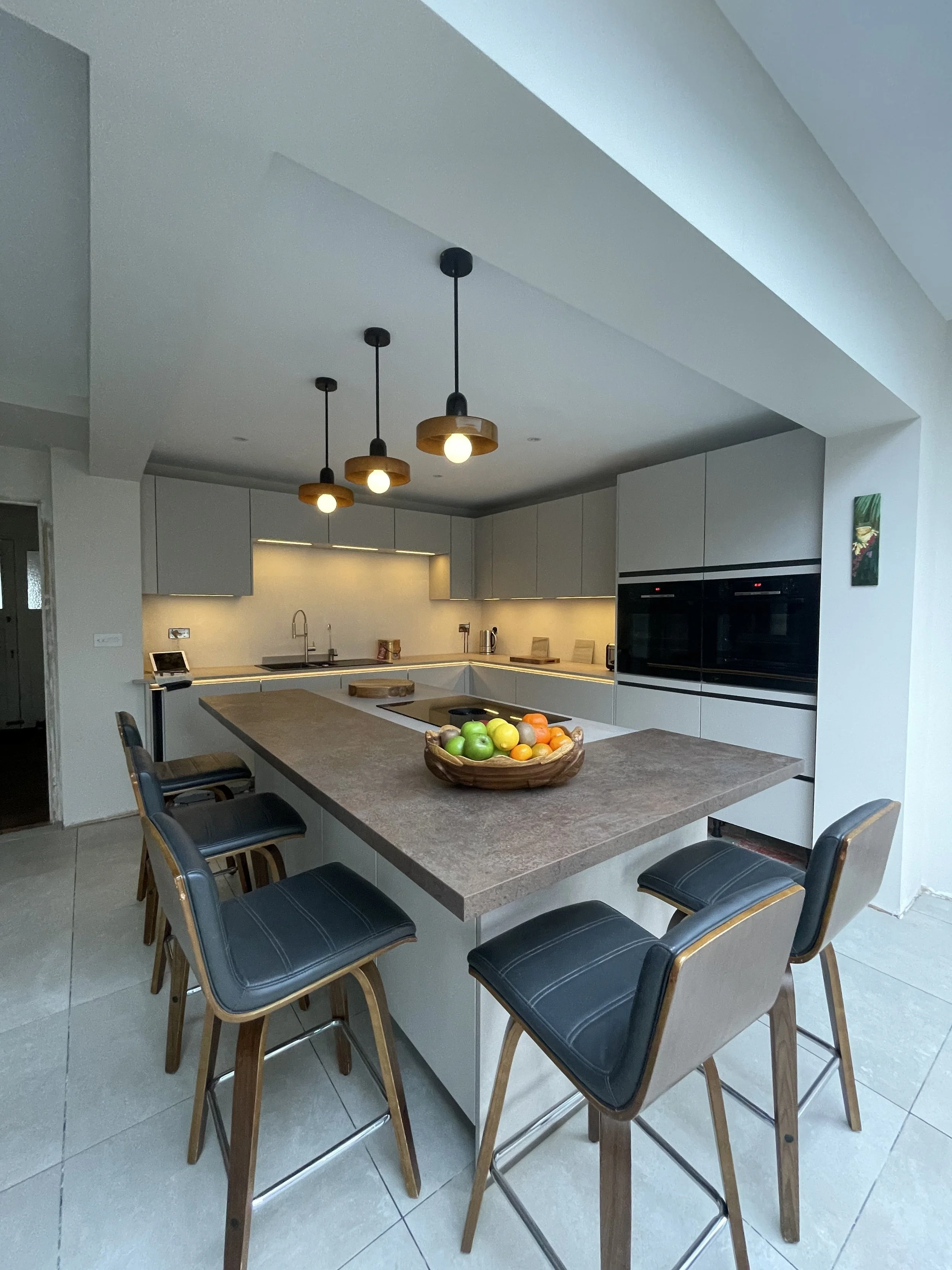 Modern kitchen with a large central island, black bar stools, pendant lights, white cabinets, and a built-in oven.