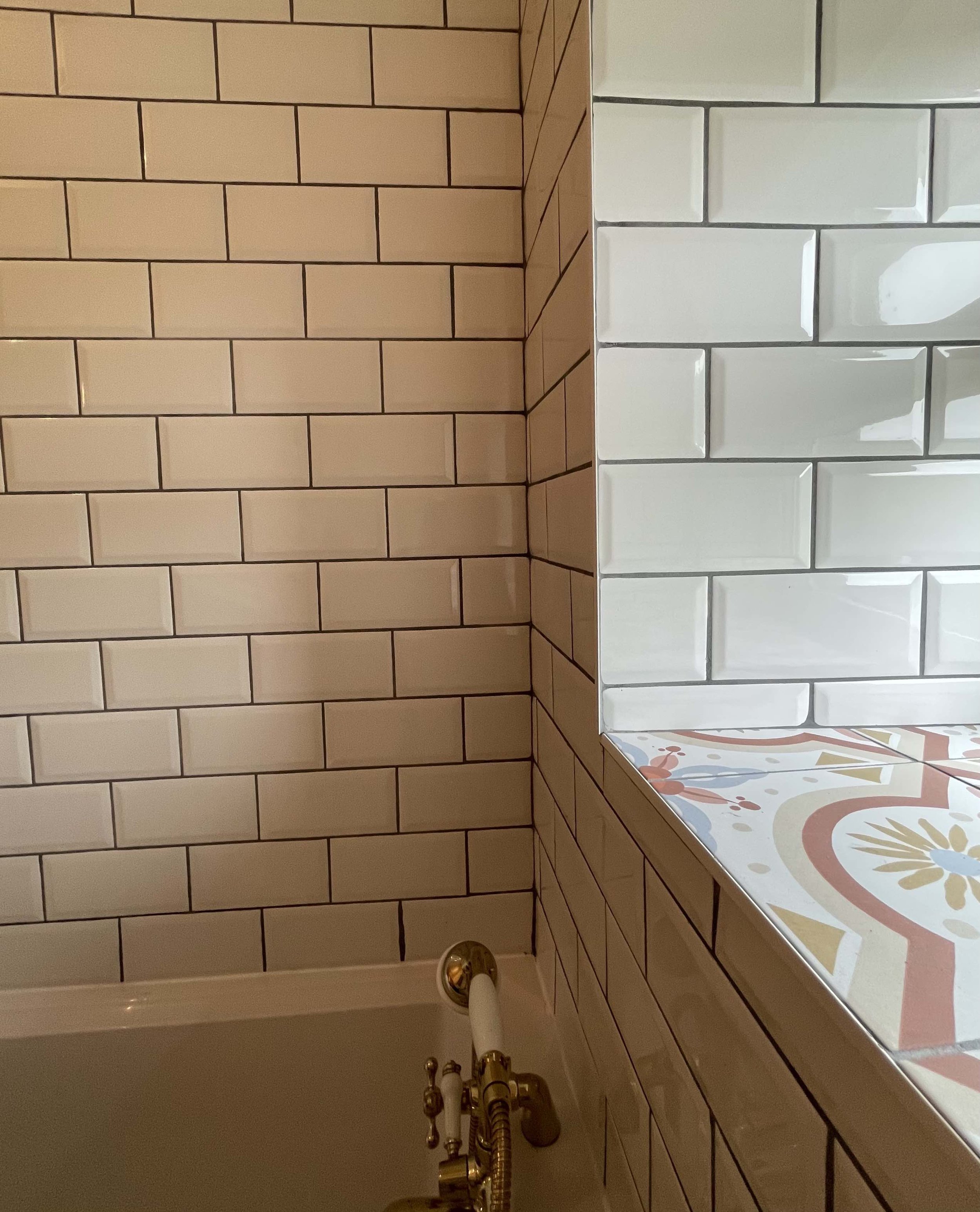Interior view of a tub area with beige and white subway tiles, a decorative floral patterned tile surface, and gold-colored fixtures.