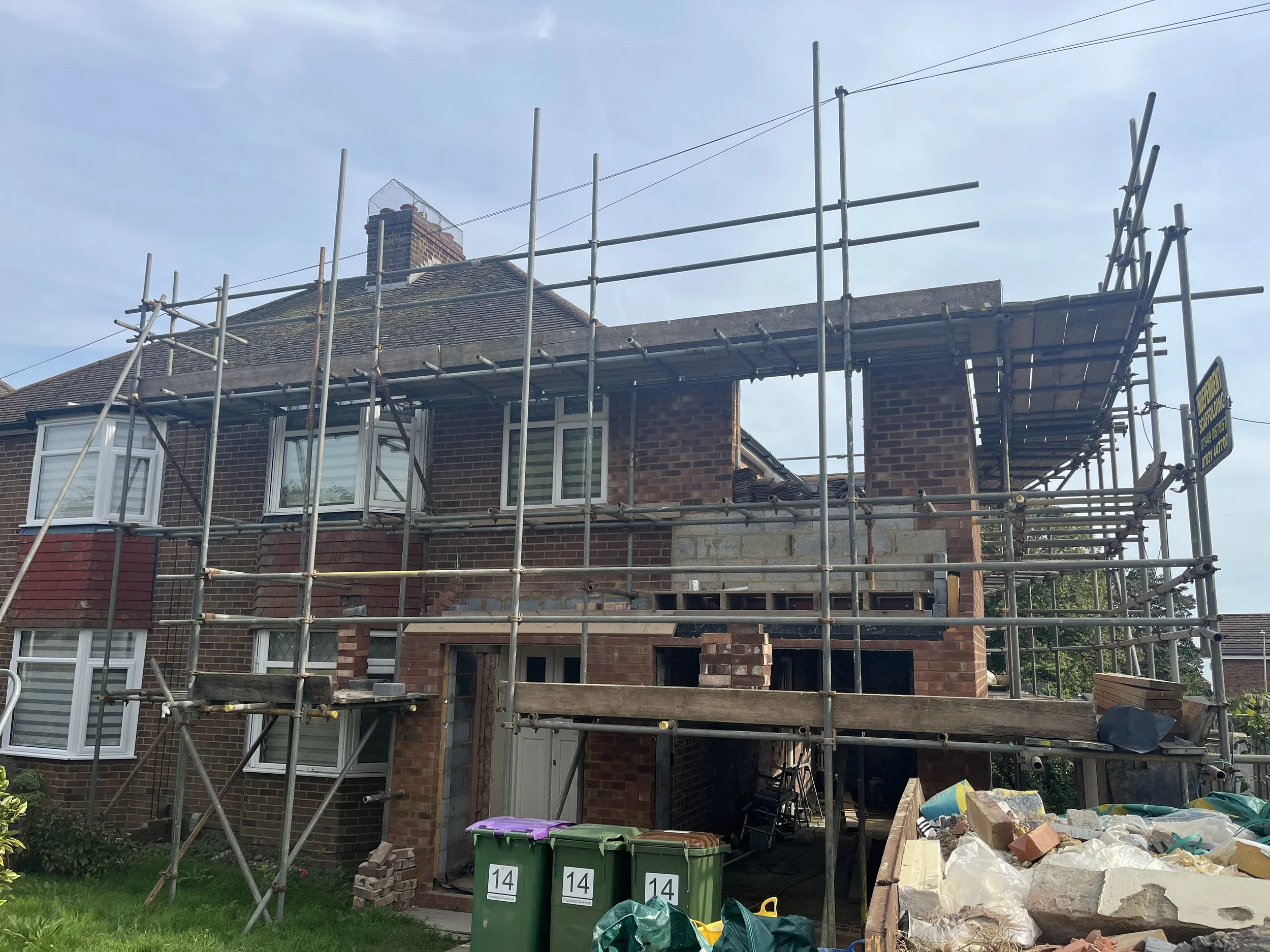 A two-story brick house under construction with scaffolding surrounding it, and construction materials and debris on the ground.