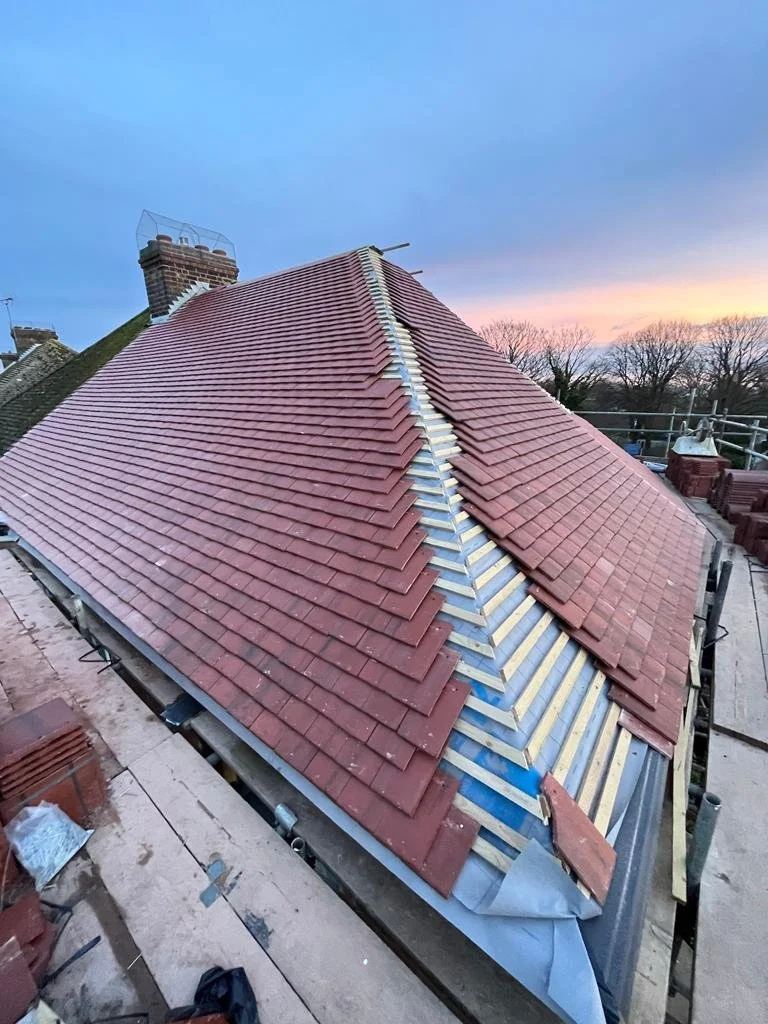 A house roof under renovation with red tiles partially installed, revealing the underlying structure and construction materials, during sunset.