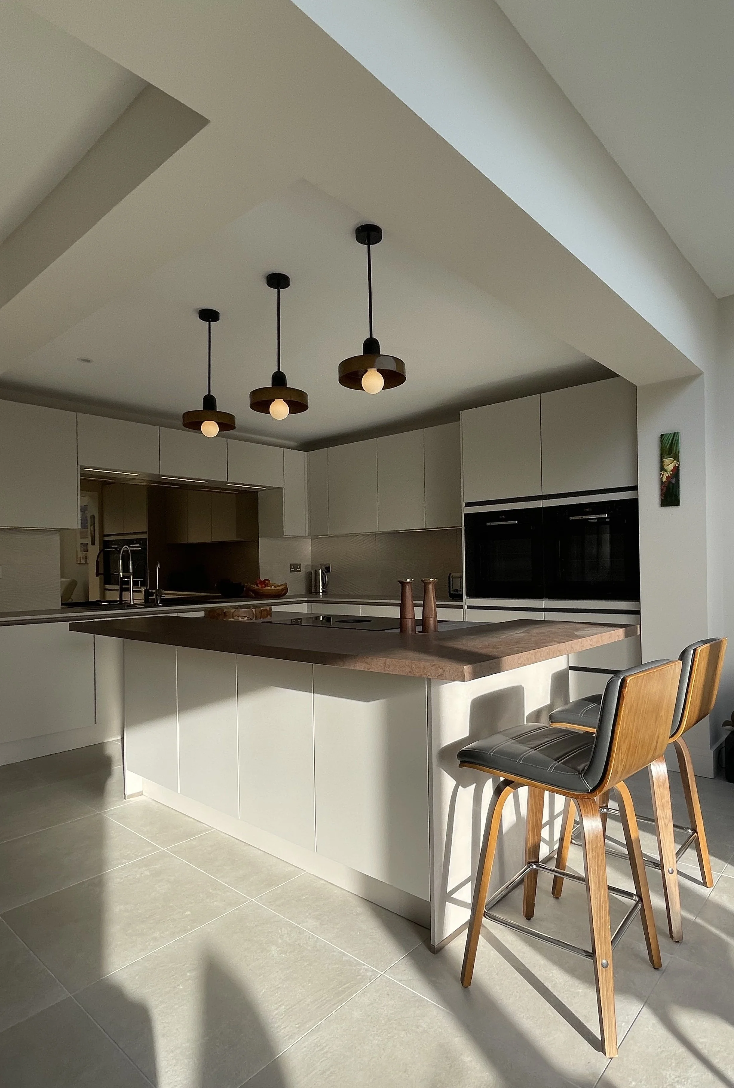 Modern kitchen with sand cabinets, a wooden countertop island, black pendant lights, and two wooden barstools with grey cushions, Kitchen Renovation Folkestone, Kent. 