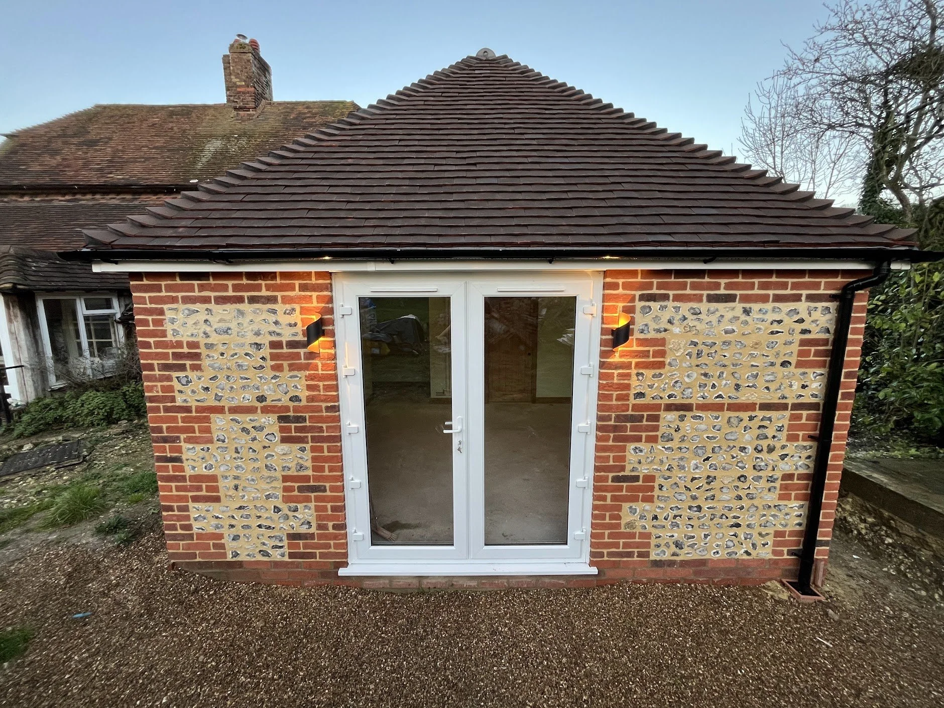 Back view of a small brick house with a pitched tile roof, white patio doors, and wall-mounted outdoor lights flanking the doors, new build in Alkham Valley, Kent. 