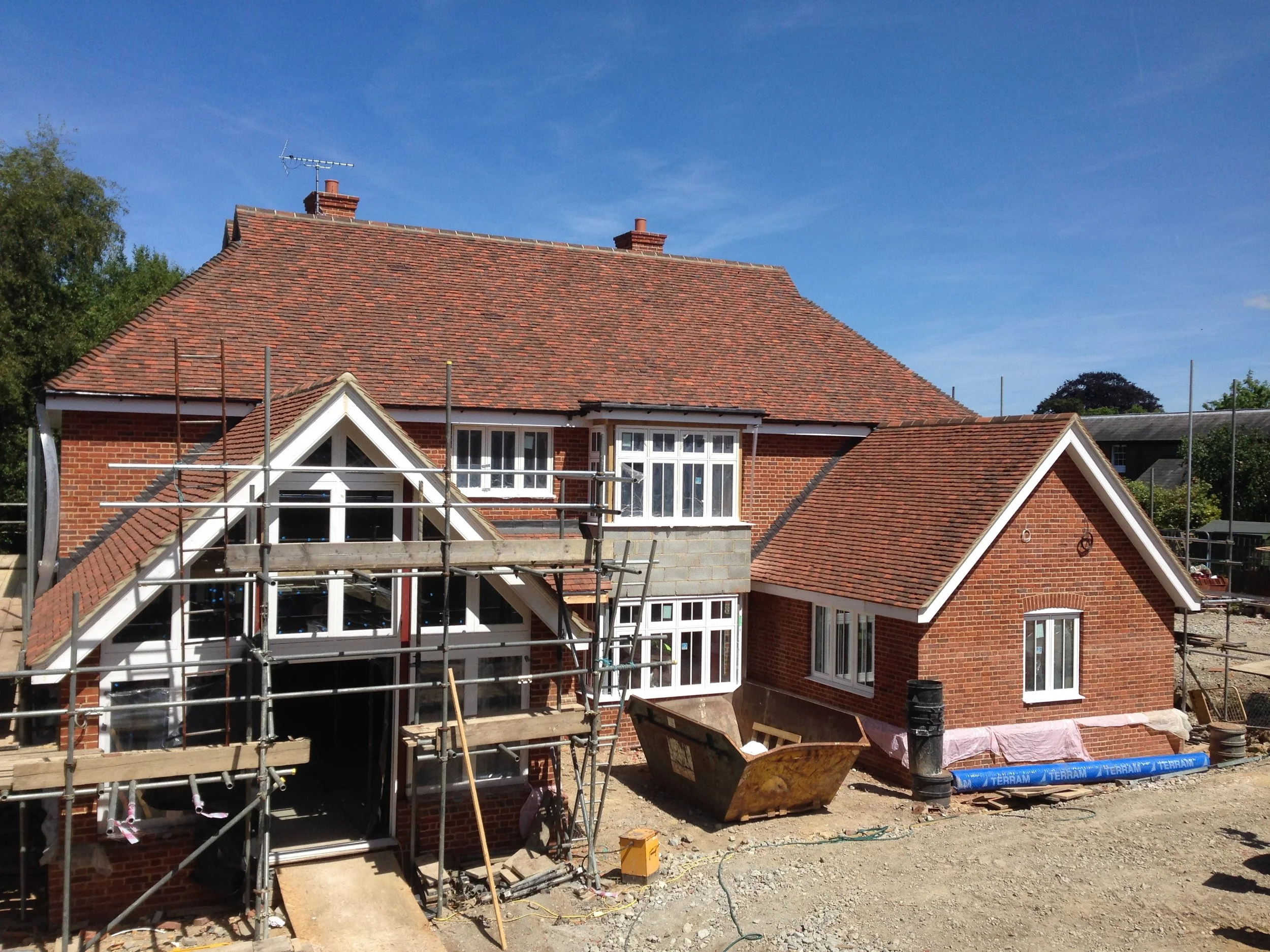 A house under construction with scaffolding, red brick walls, a steep roof with red tiles, and multiple windows, including a prominent front window in a gable.
