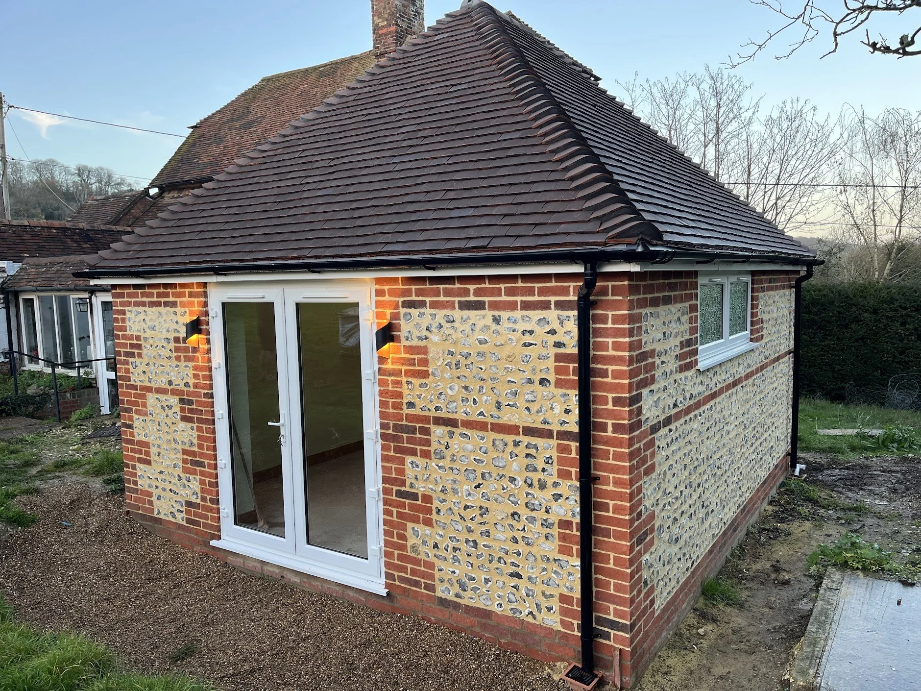 Small brick and stone house, flint walls with a sloped tiled roof, white double glass patio doors, and a nearby window, surrounded by a yard.