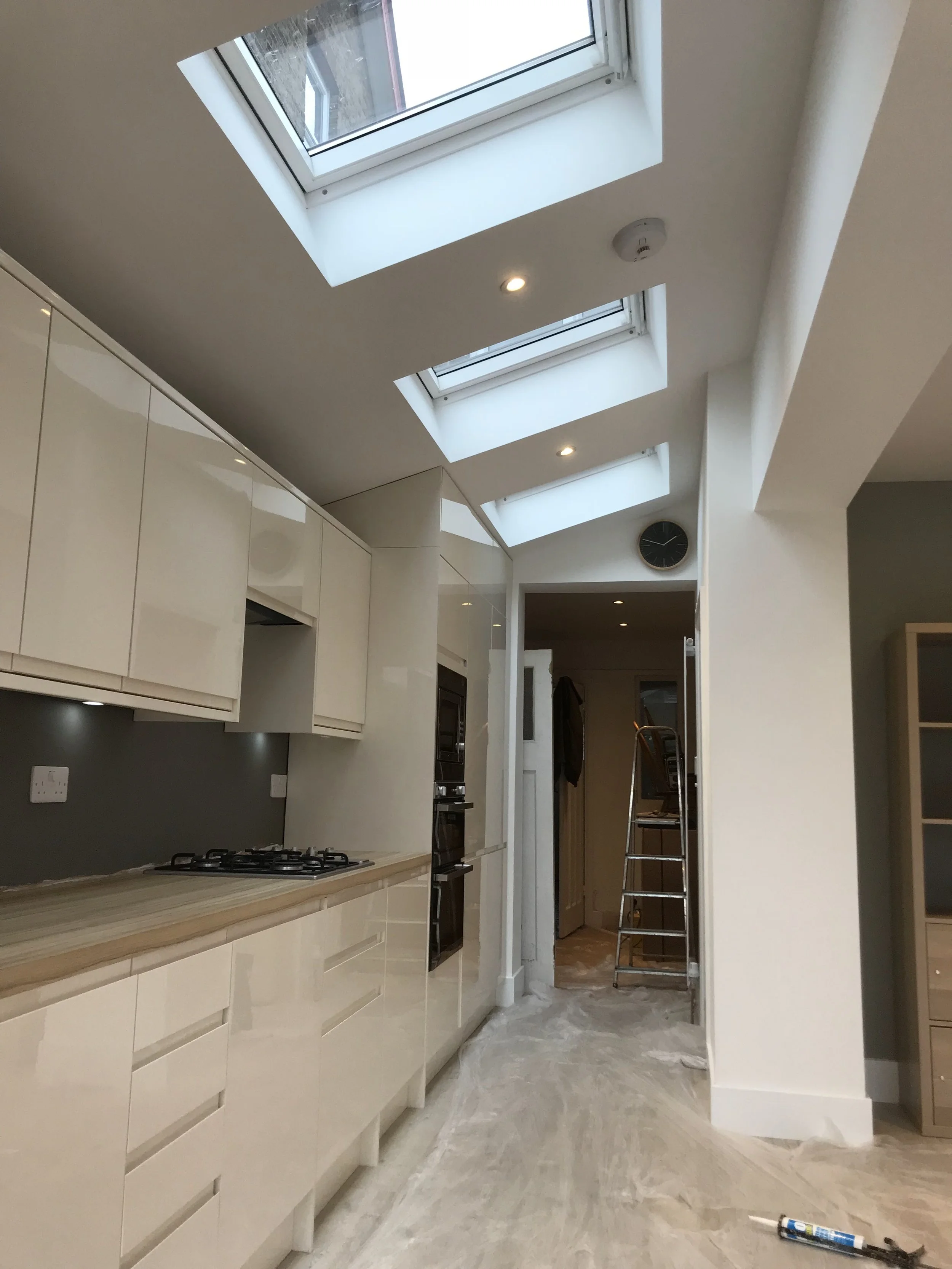 Kitchen with white cabinetry, wooden countertop, and three skylights in the ceiling. There are built-in ovens, a hanging clock, a ladder, and a doorway leading to another room. Construction materials are on the floor.