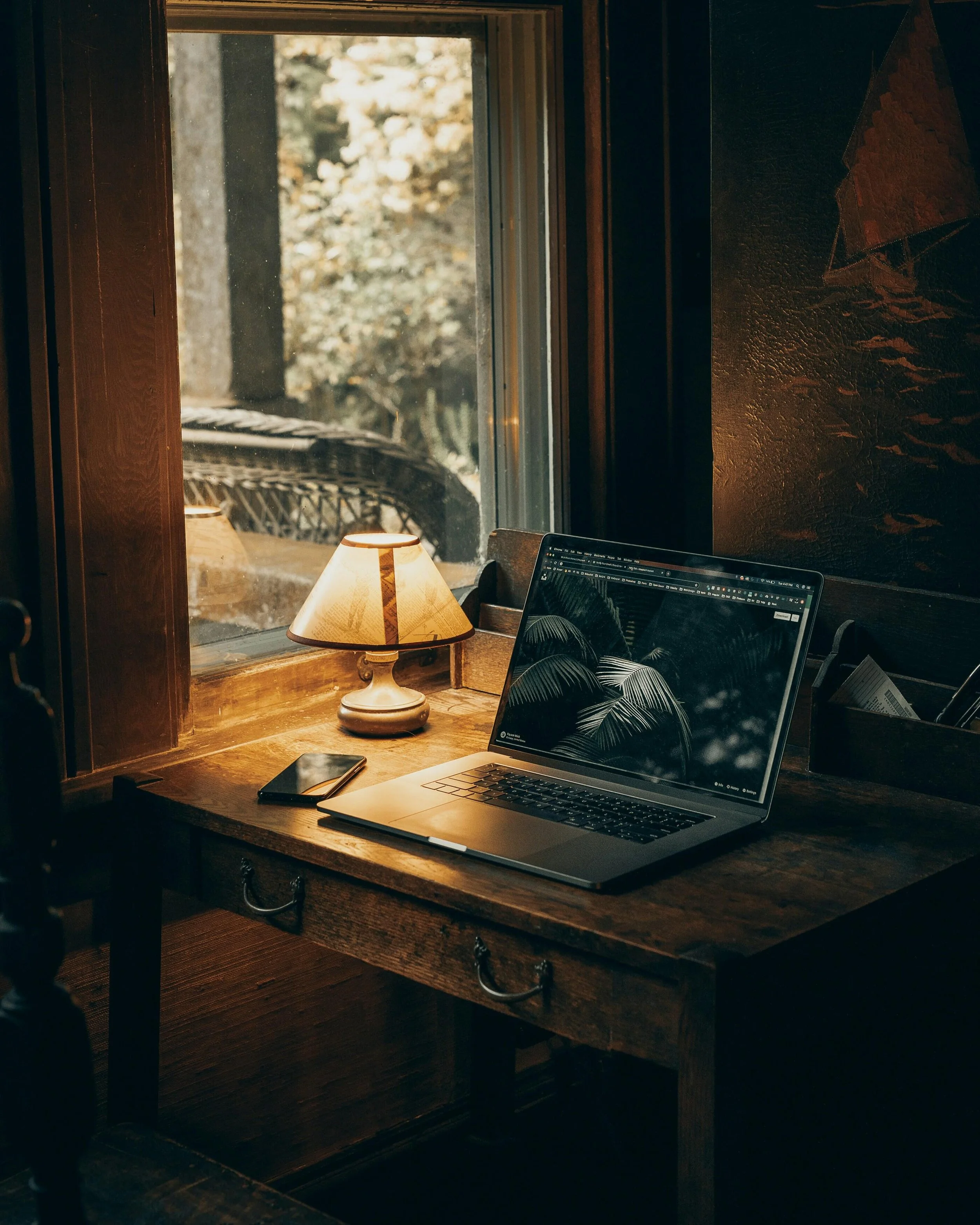 A wooden desk beside a window with a lamp, smartphone, and open laptop displaying a dark leaf pattern, in a cozy, dimly lit room.